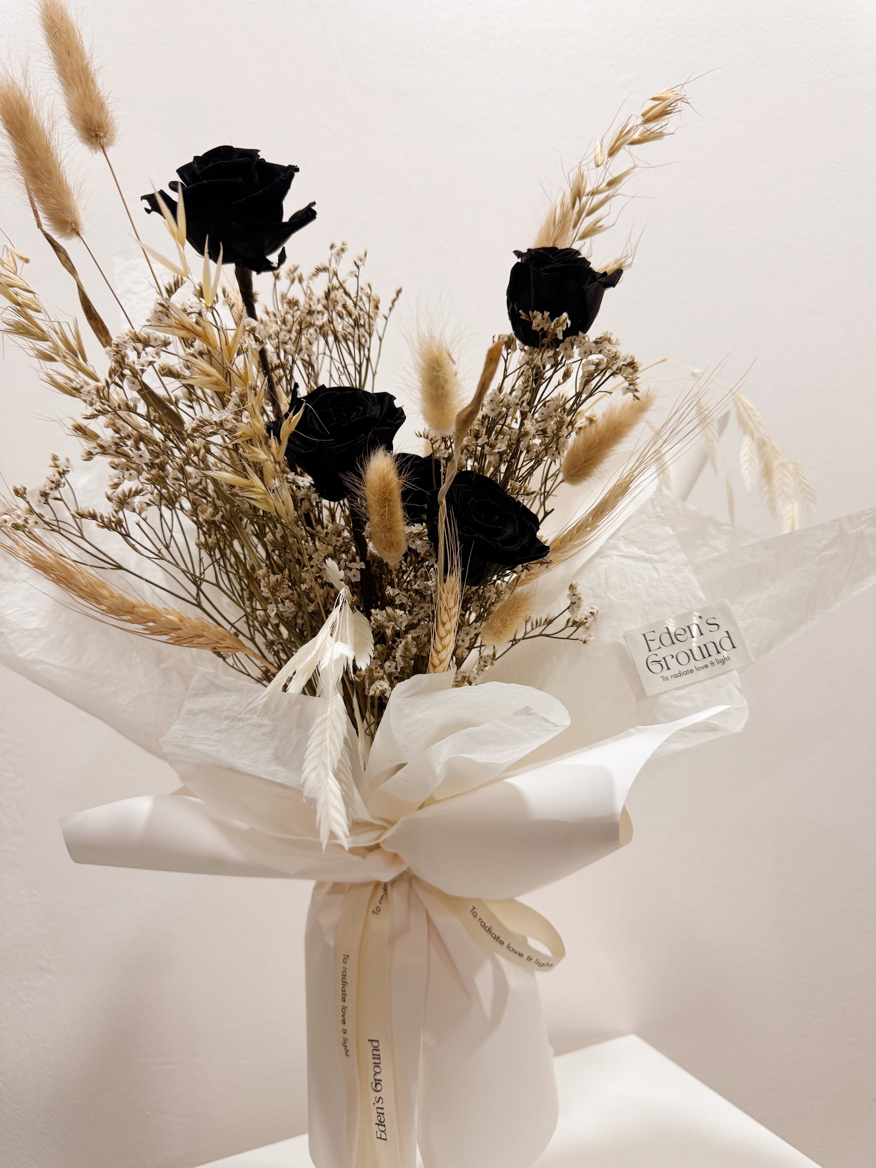 Noir Muse bouquet made with preserved black roses along with filler flowers and foliage, wrapped in white paper crafted by Eden’s Ground, a floral design boutique.