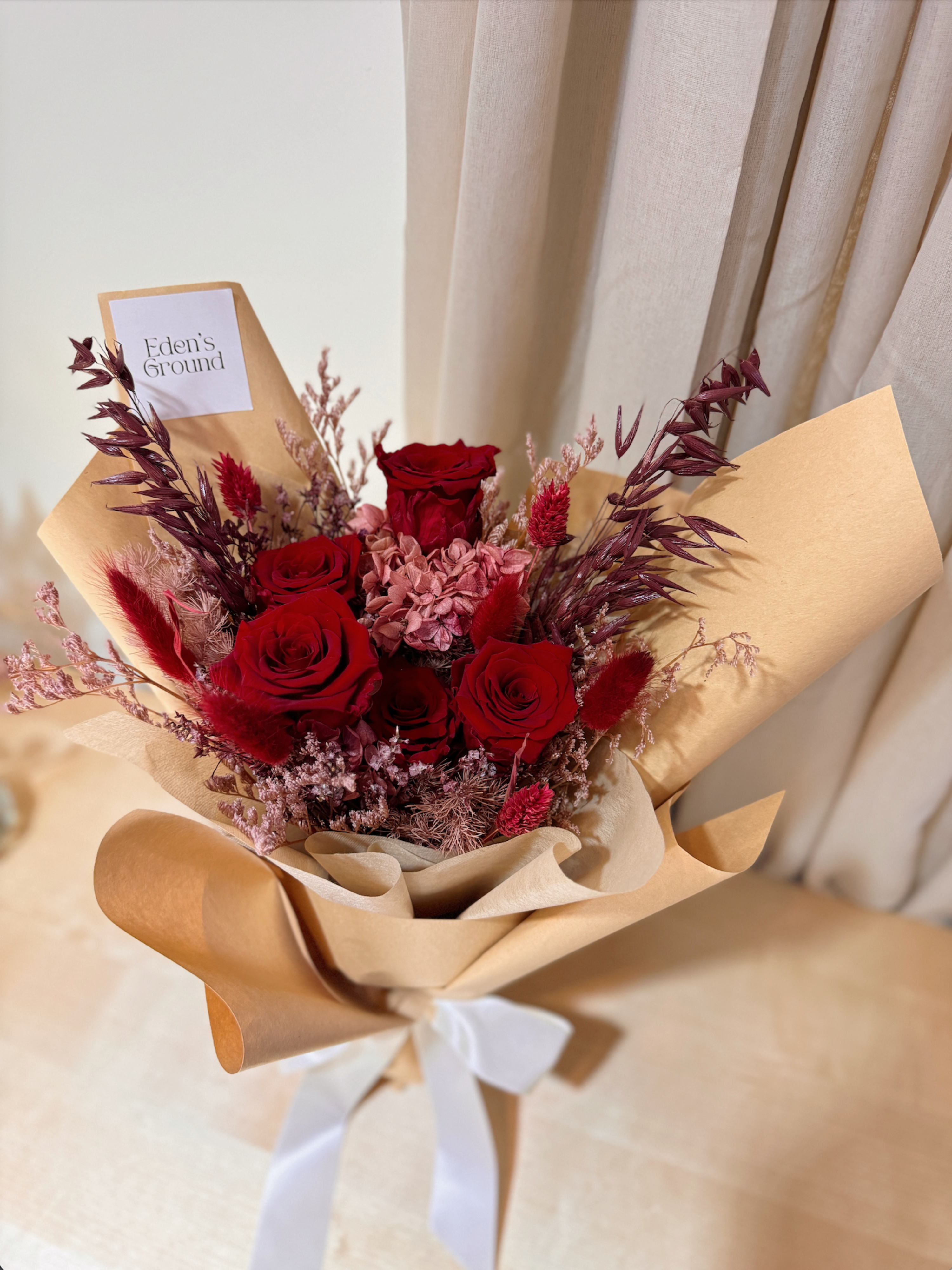 Eternal valentine bouquet made with all shades of red to symbolise deep love, surrounded by textural foliage, wrapped in tissue paper and kraft paper, crafted by Eden’s Ground.