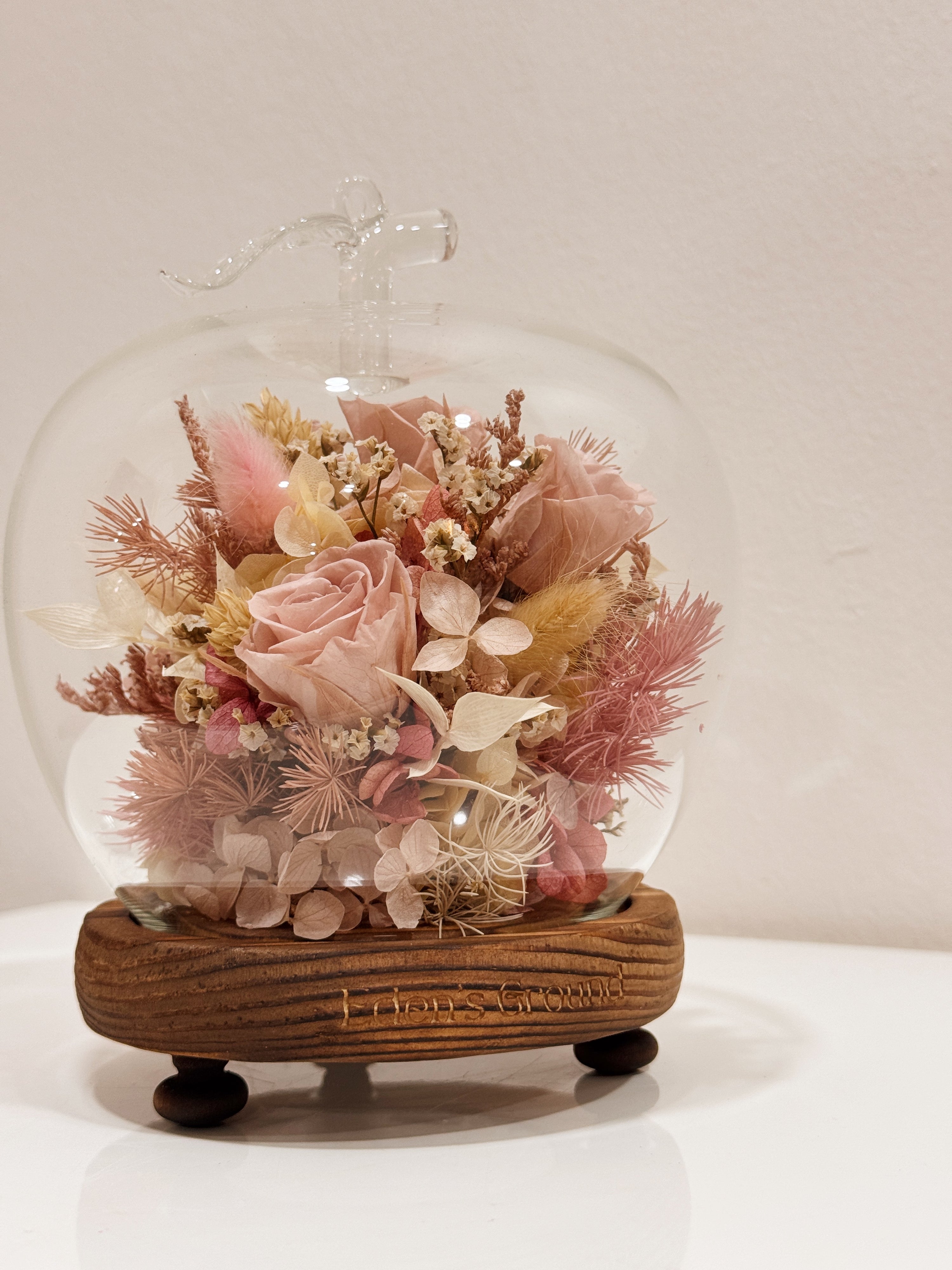 A delicate blend of preserved and dried flowers in soft pink tones, beautifully arranged in a lighted apple-shaped glass dome by Eden’s Ground—an enchanting keepsake made to last.