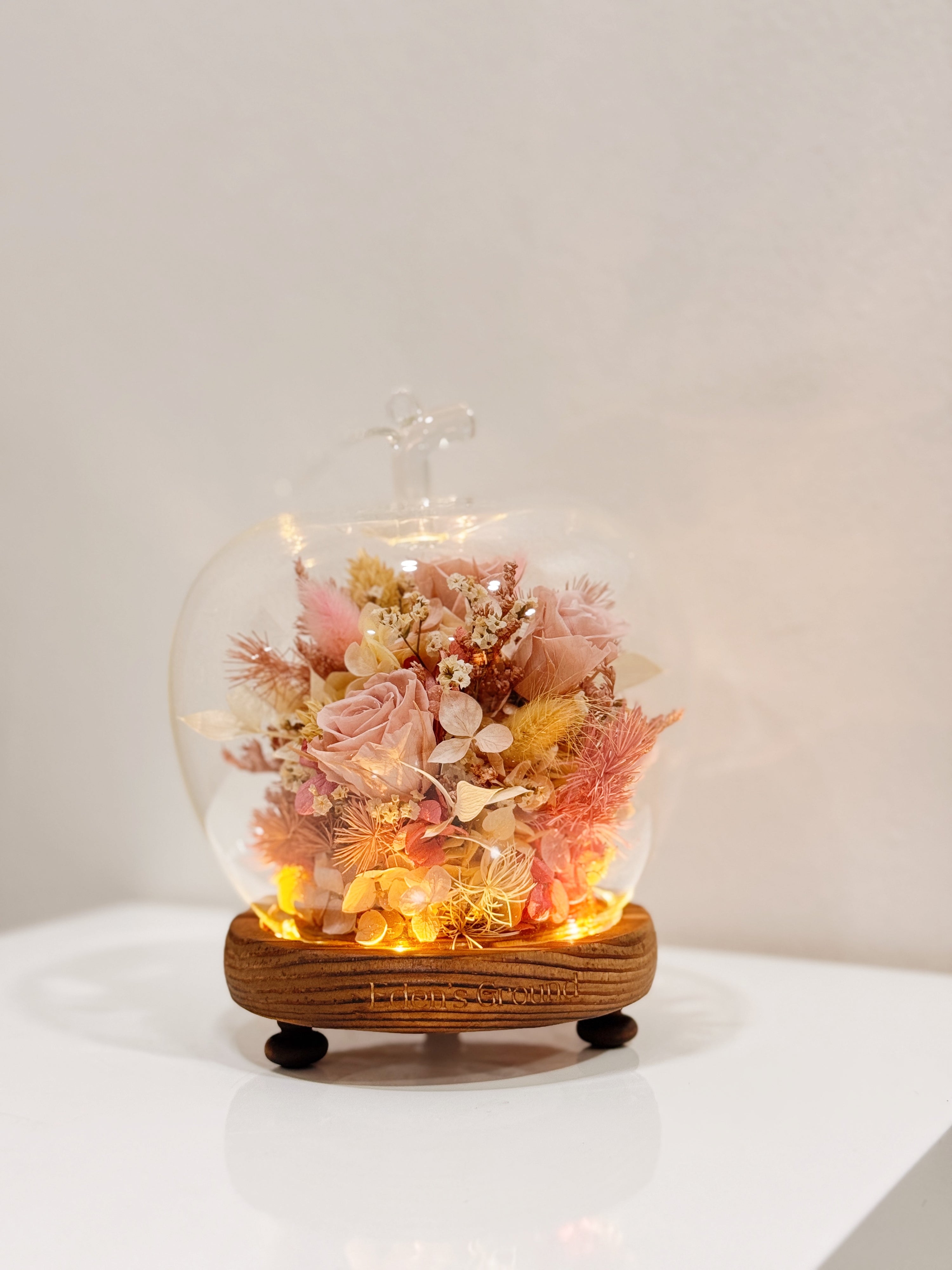 A delicate blend of preserved and dried flowers in soft pink tones, beautifully arranged in a lighted apple-shaped glass dome by Eden’s Ground—an enchanting keepsake made to last. 