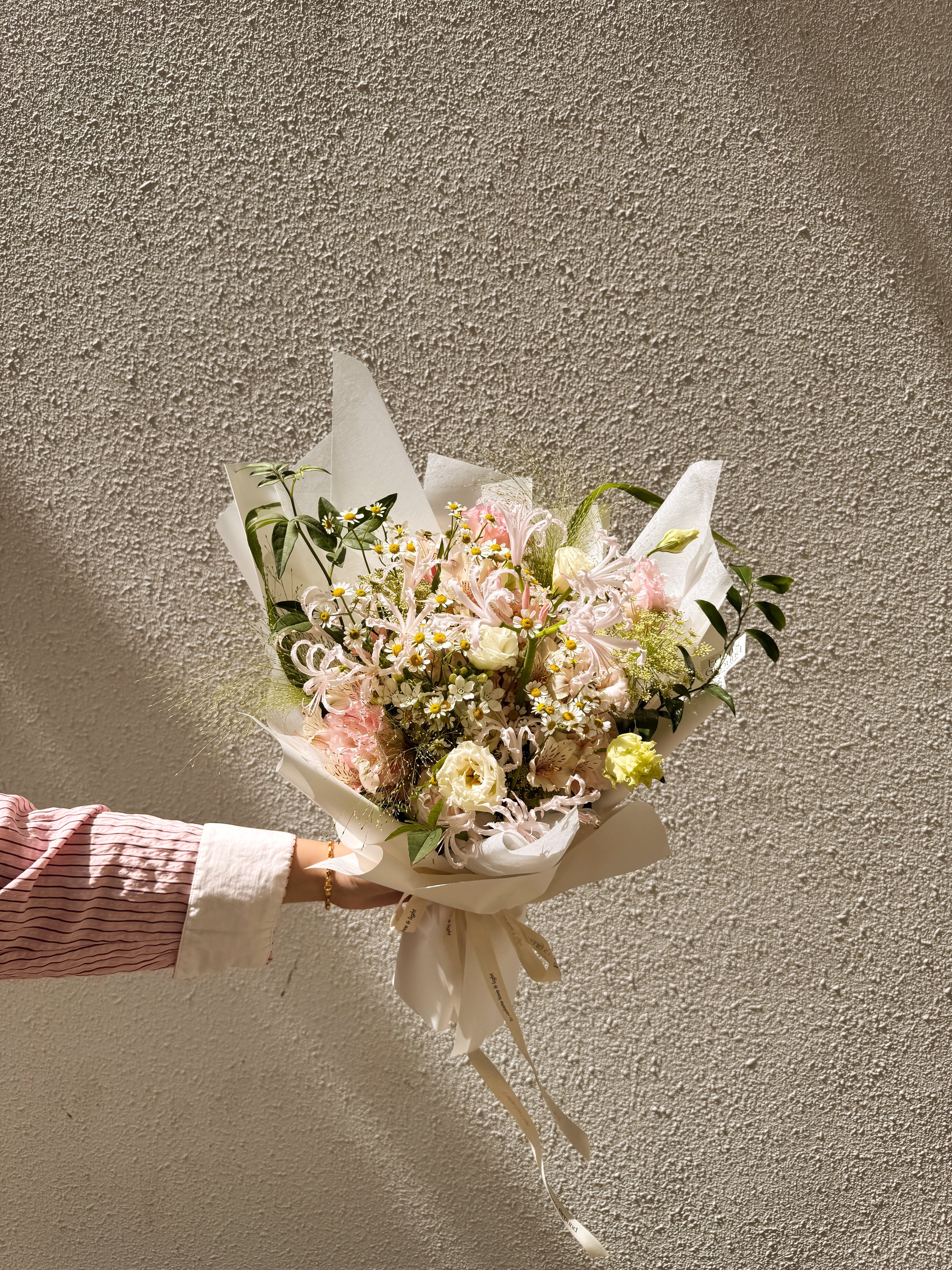 Pink flowers with daisy mix Bouquet, along with filler foliage, wrapped with white tissue paper and white glassine paper, crafted by Eden's Ground, a floral design boutique.
