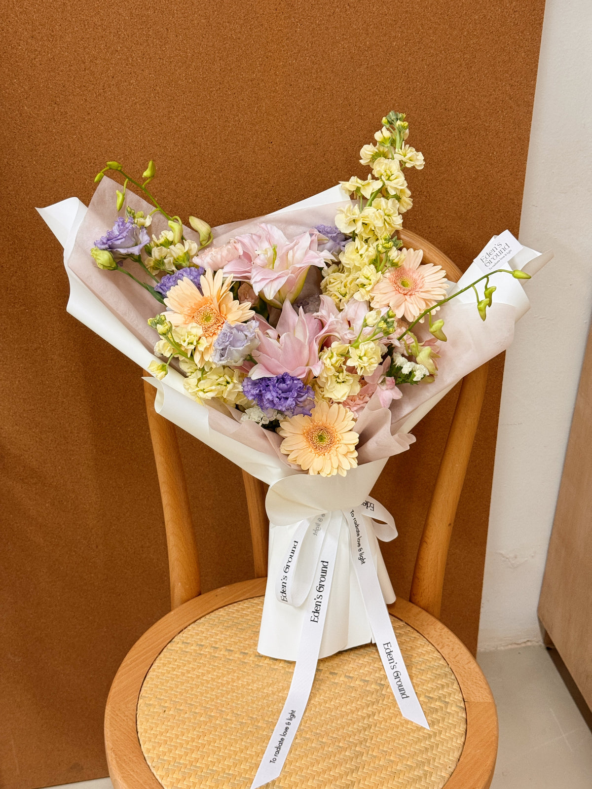 A mixed of pastel flower bouquet, wrapped in white korean paper and pink tissue paper lining. The bouquet features light pink rose lily and peach daisy, orchid, and delicate filler flowers. Freshly curated by Eden's Ground floral design studio in Singapore.