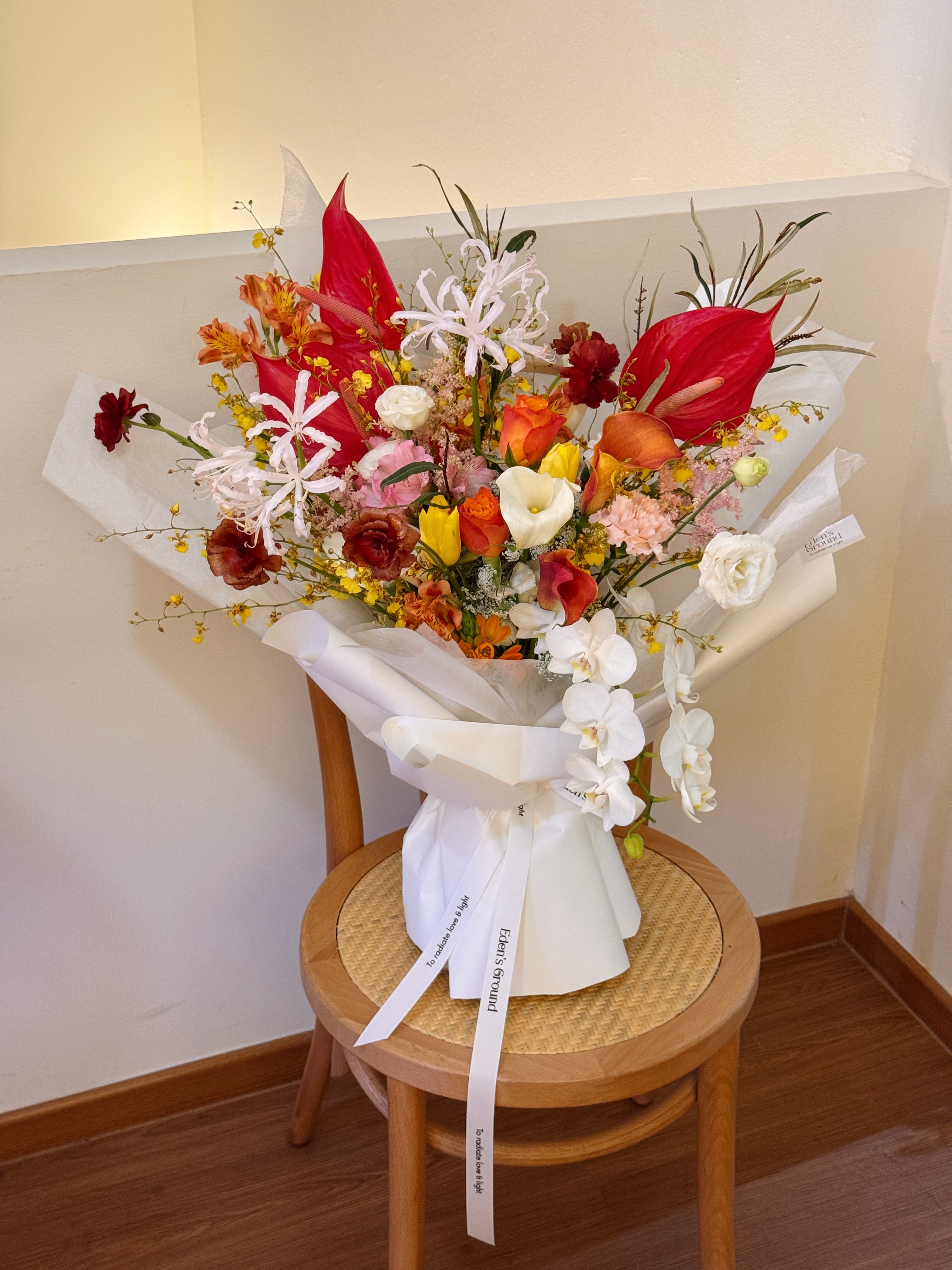 Bright pop of colour flowers with dancing lady orchid, red anthurium, butterfly ranunculus, calla lily, tulips, roses bouquet, along with filler foliage, wrapped with white tissue paper and white glassine paper, crafted by Eden's Ground, a floral design boutique.