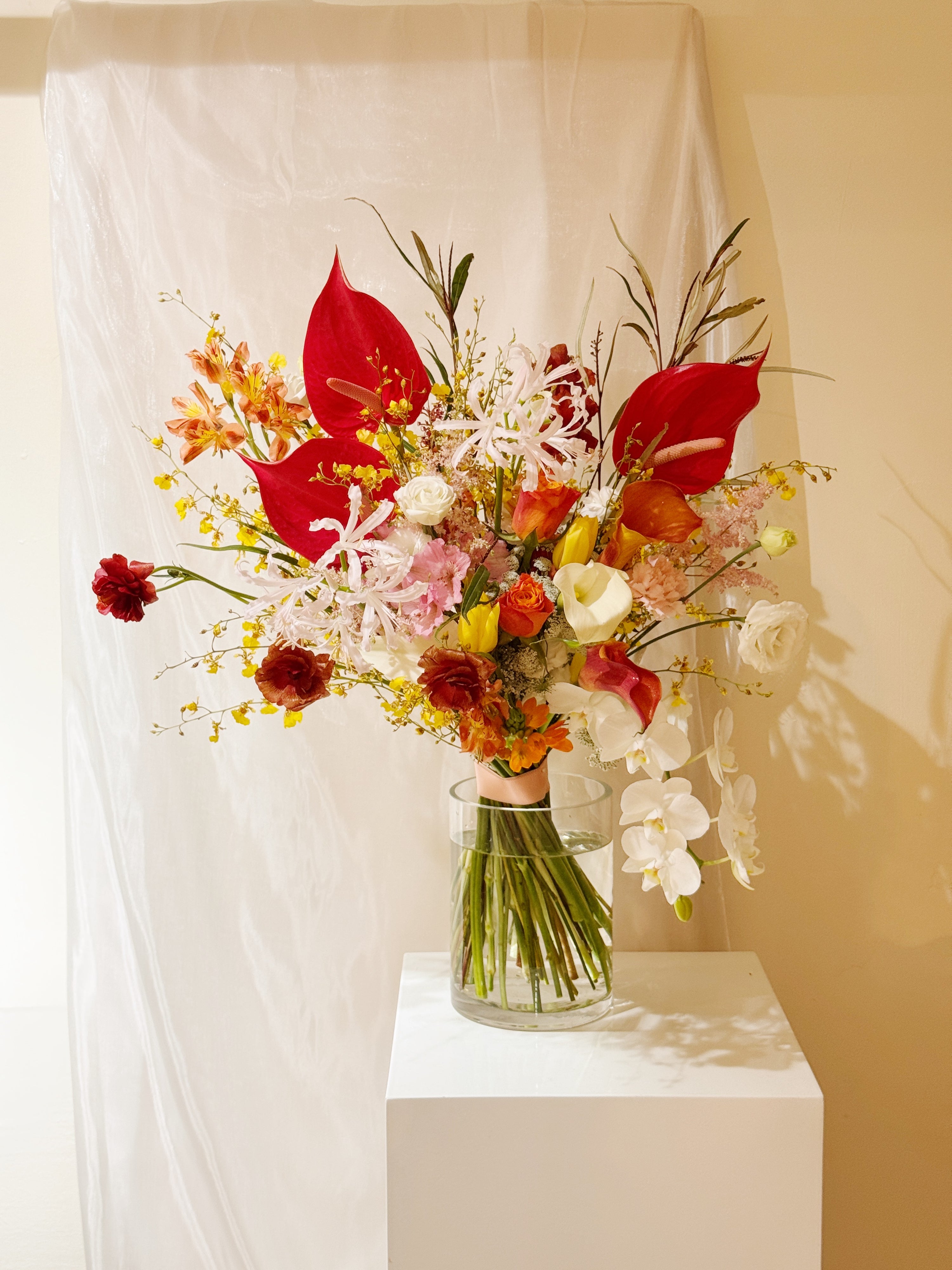 Bright pop of colour flowers with dancing lady orchid, red anthurium, butterfly ranunculus, calla lily, tulips, roses vase arrangement, along with filler foliage, wrapped with white tissue paper and white glassine paper, crafted by Eden's Ground, a floral design boutique.