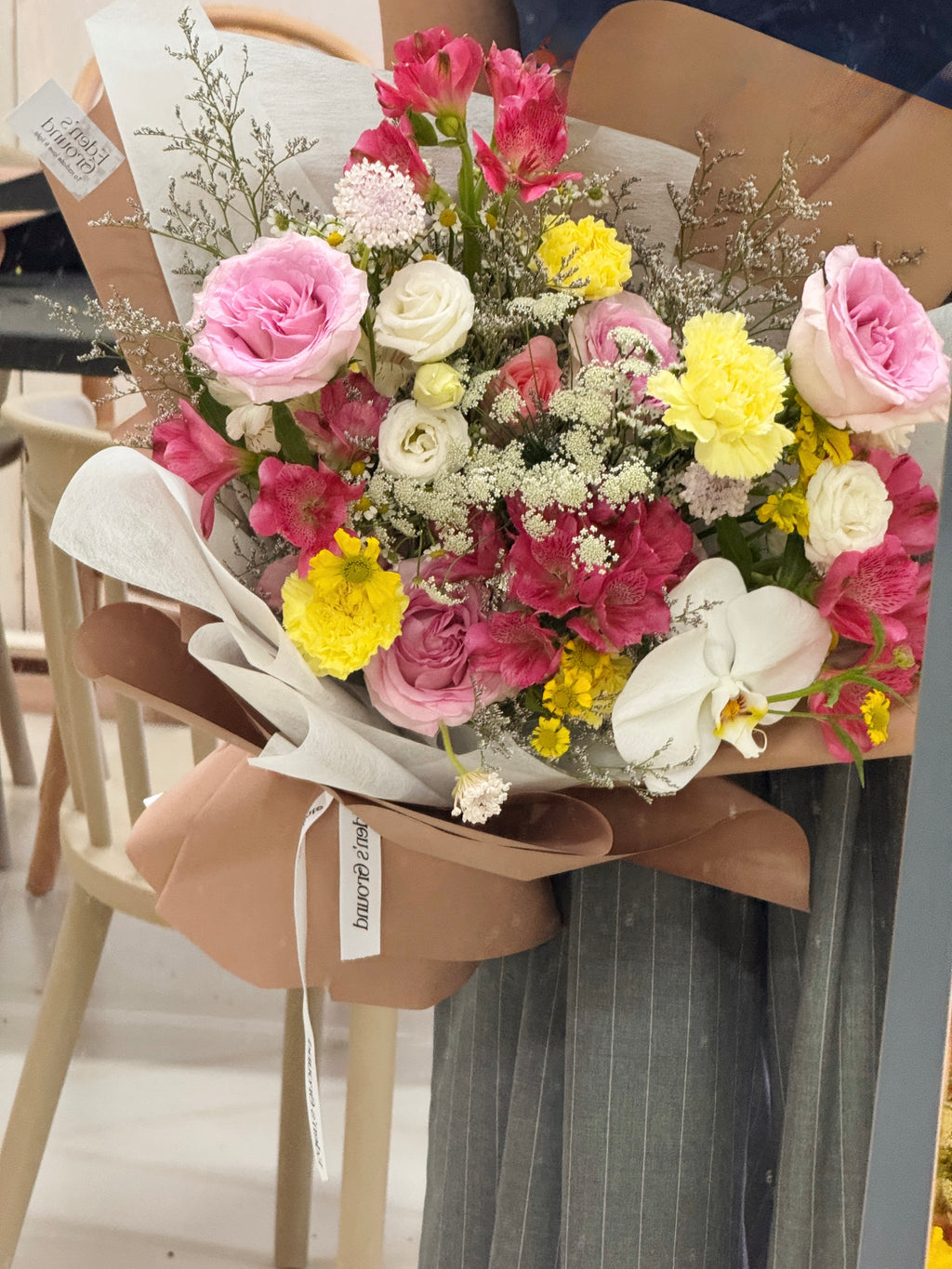 A vibrant mixed flower bouquet, wrapped in brown kraft paper and white tissue paper. The bouquet features pink and white roses, yellow daisies, pink carnations, white orchids, and delicate filler flowers. Freshly curated by Eden's Ground floral design studio in Singapore.