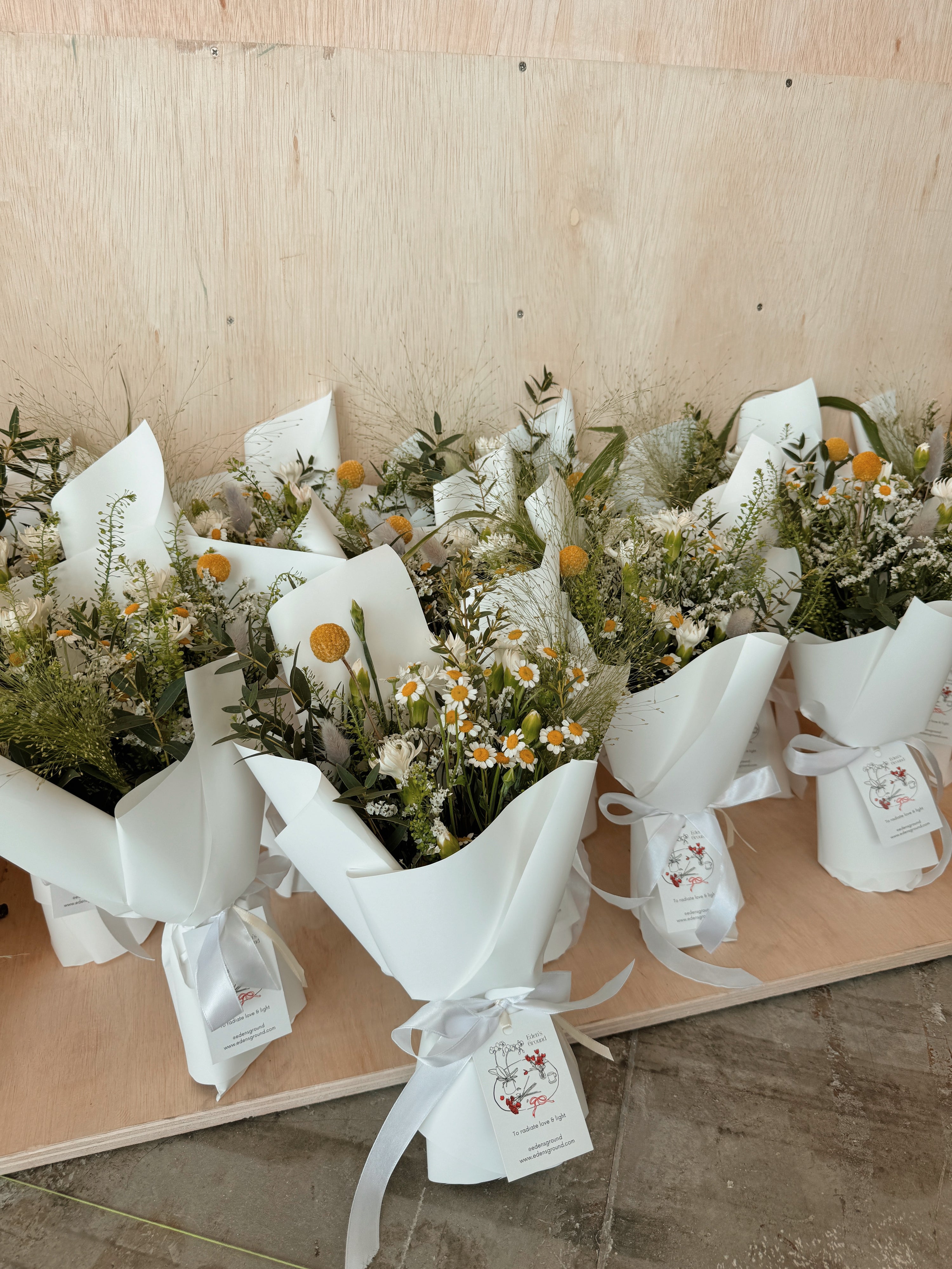 Blooms in the morning light bouquet featuring nature-inspired wildflowers with chamomile daisy, filled with lots of textural foliage such as eucalyptus, wrapped in white tissue paper and white glassine paper crafted by Eden’s Ground.
