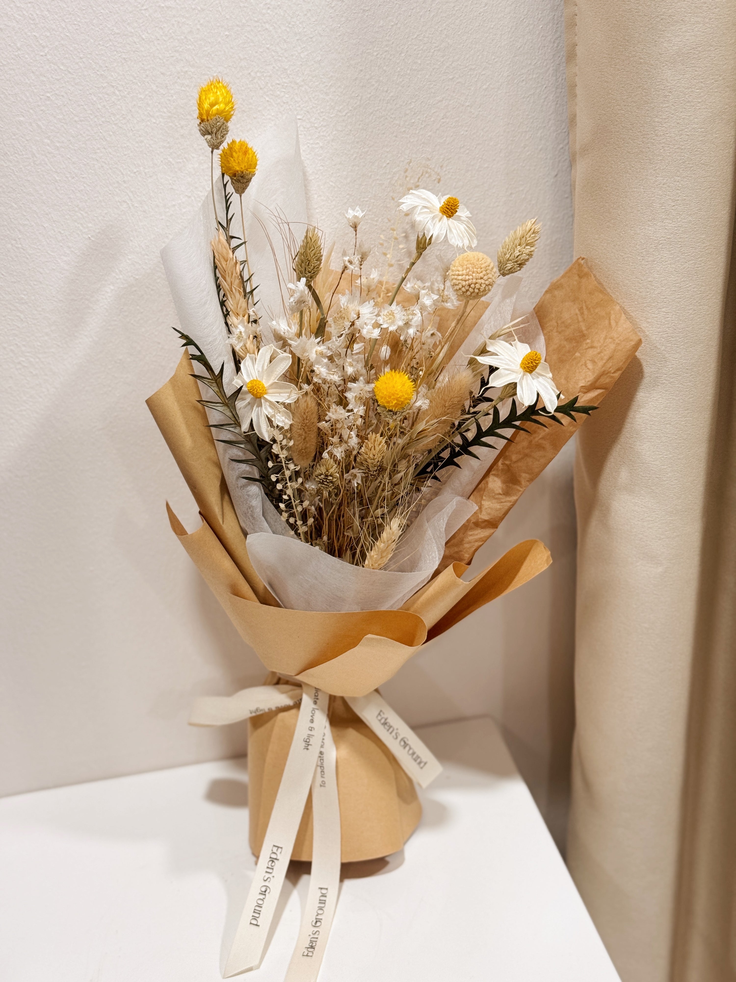 Bouquet of dried daisies with white and yellow filler flower, wrapped with brown Kraft paper, crafted by Eden's Ground.