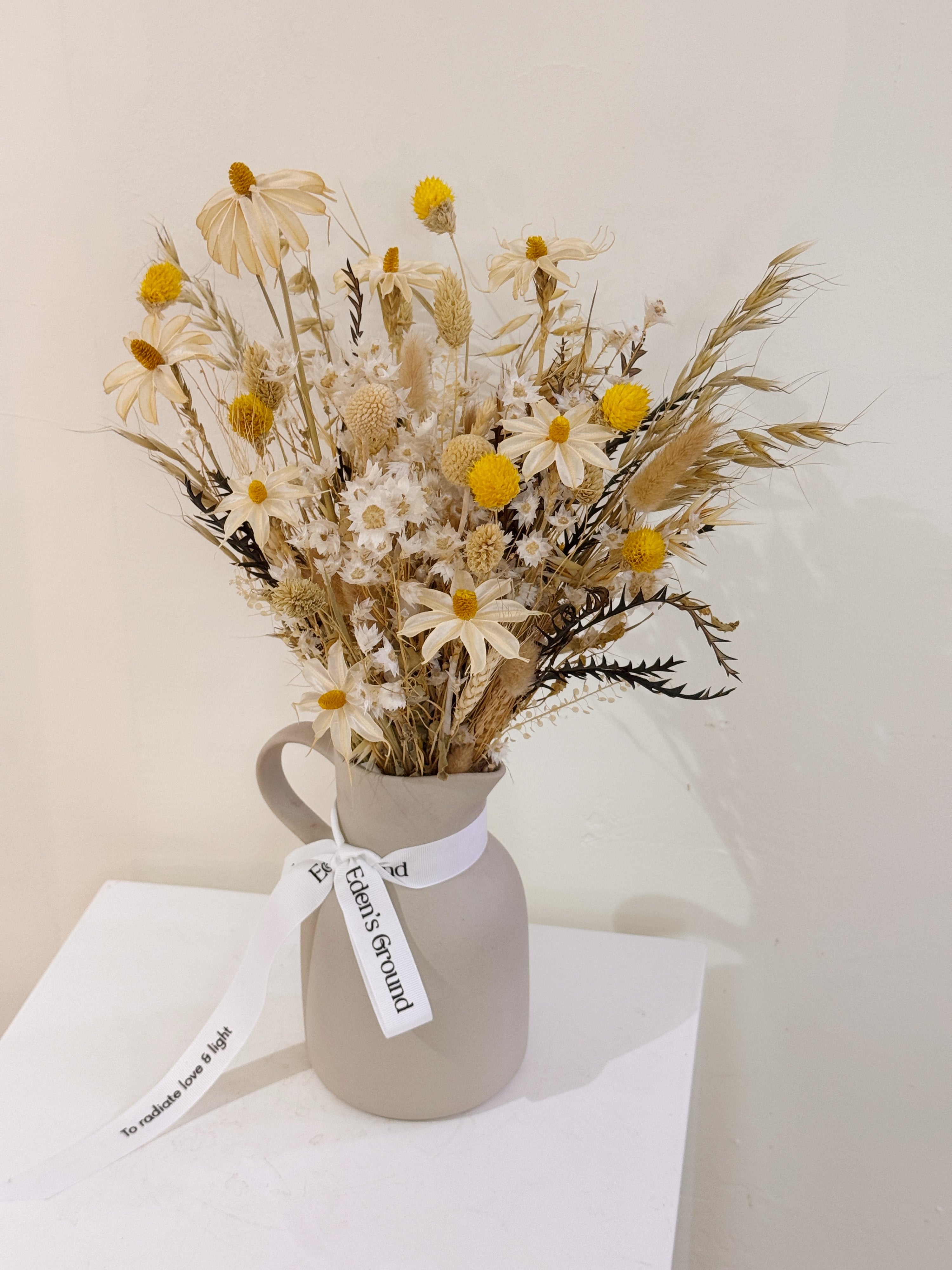 Jug arrangement of dried daisies with white and yellow filler flower, arranged in a clear glass vase, crafted by Eden's Ground.