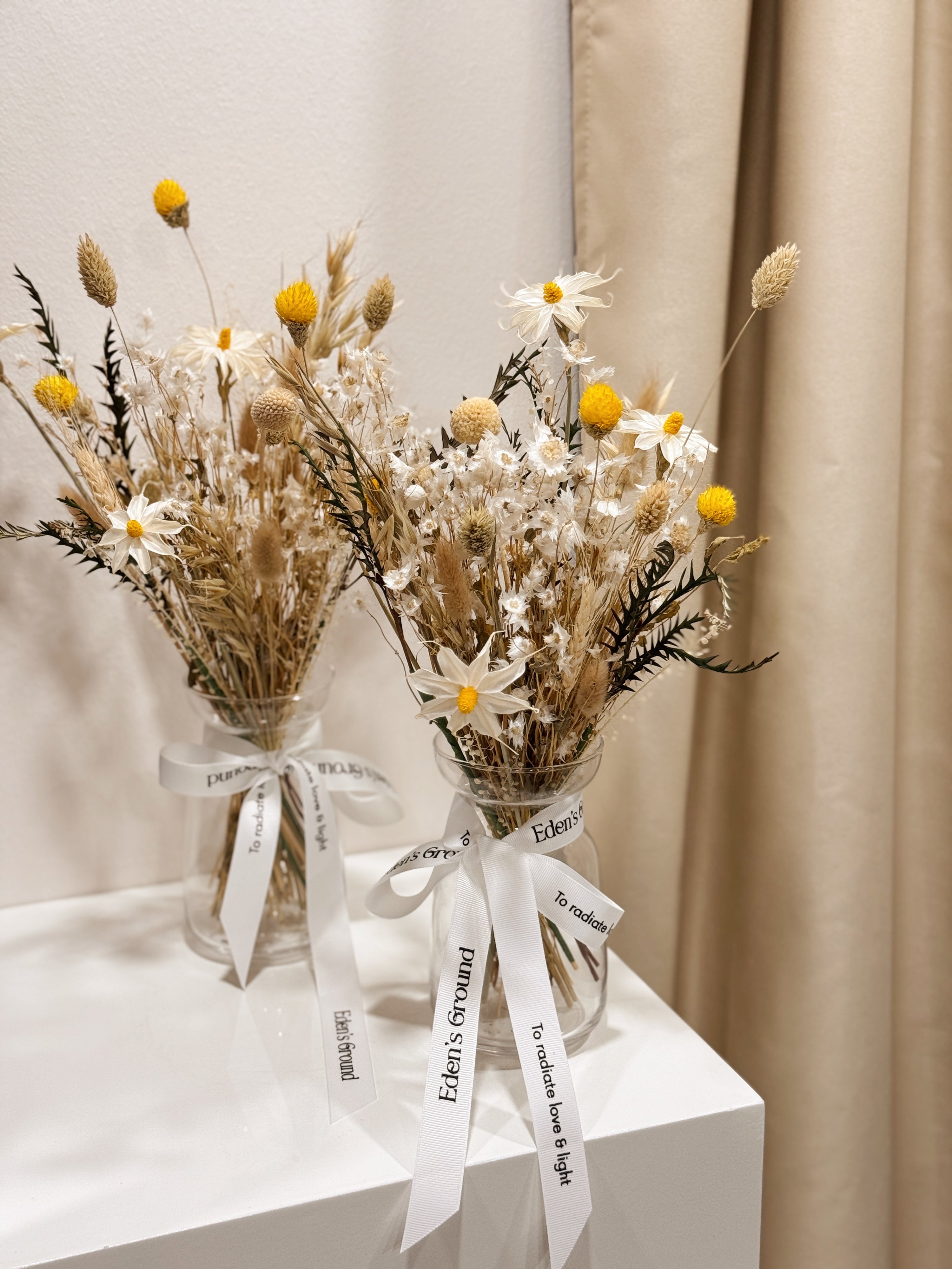 Vase arrangement of dried daisies with white and yellow filler flower, arranged in a clear glass vase, crafted by Eden's Ground.