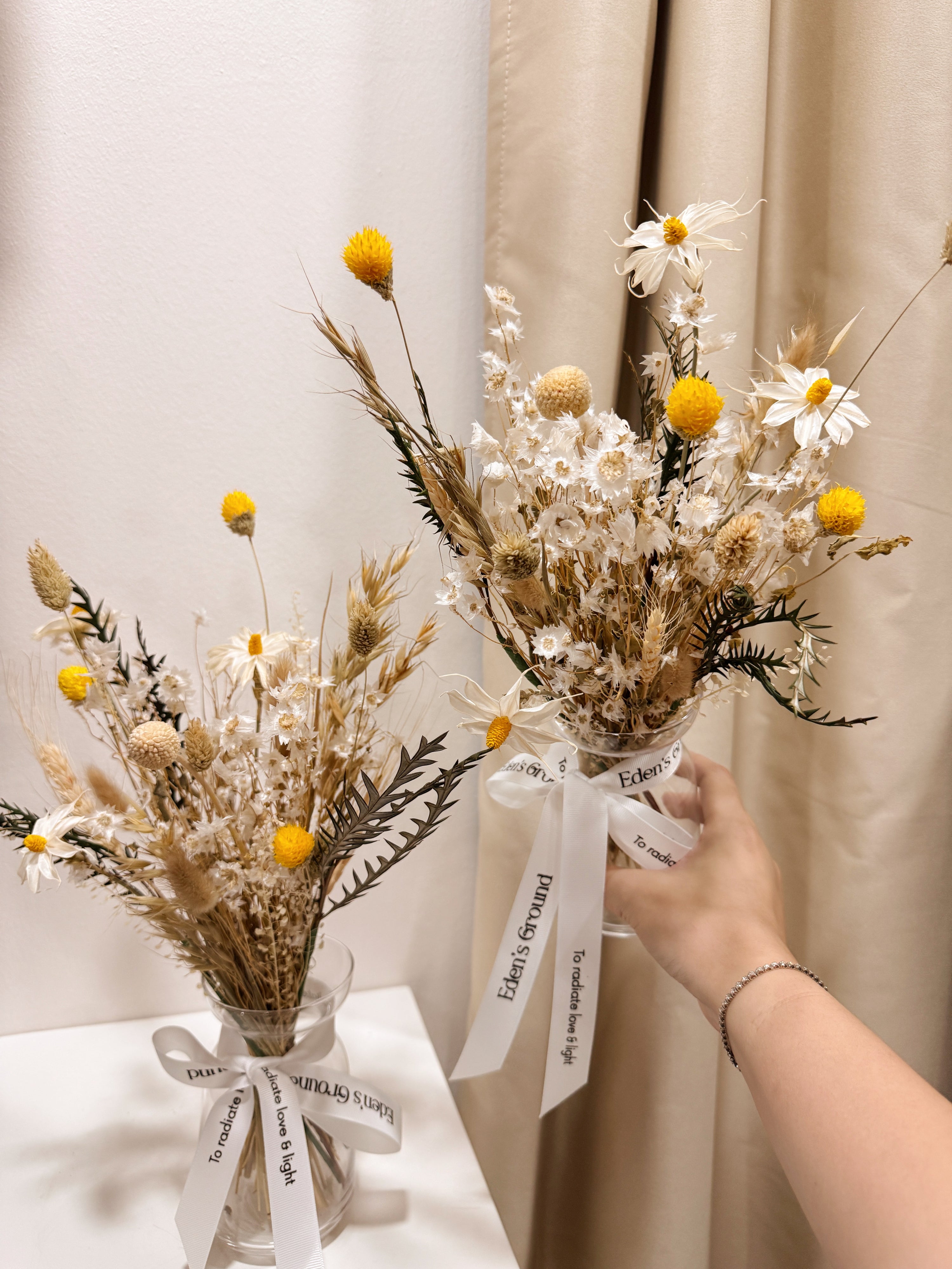 Vase arrangement of dried daisies with white and yellow filler flower, arranged in a clear glass vase, crafted by Eden's Ground.