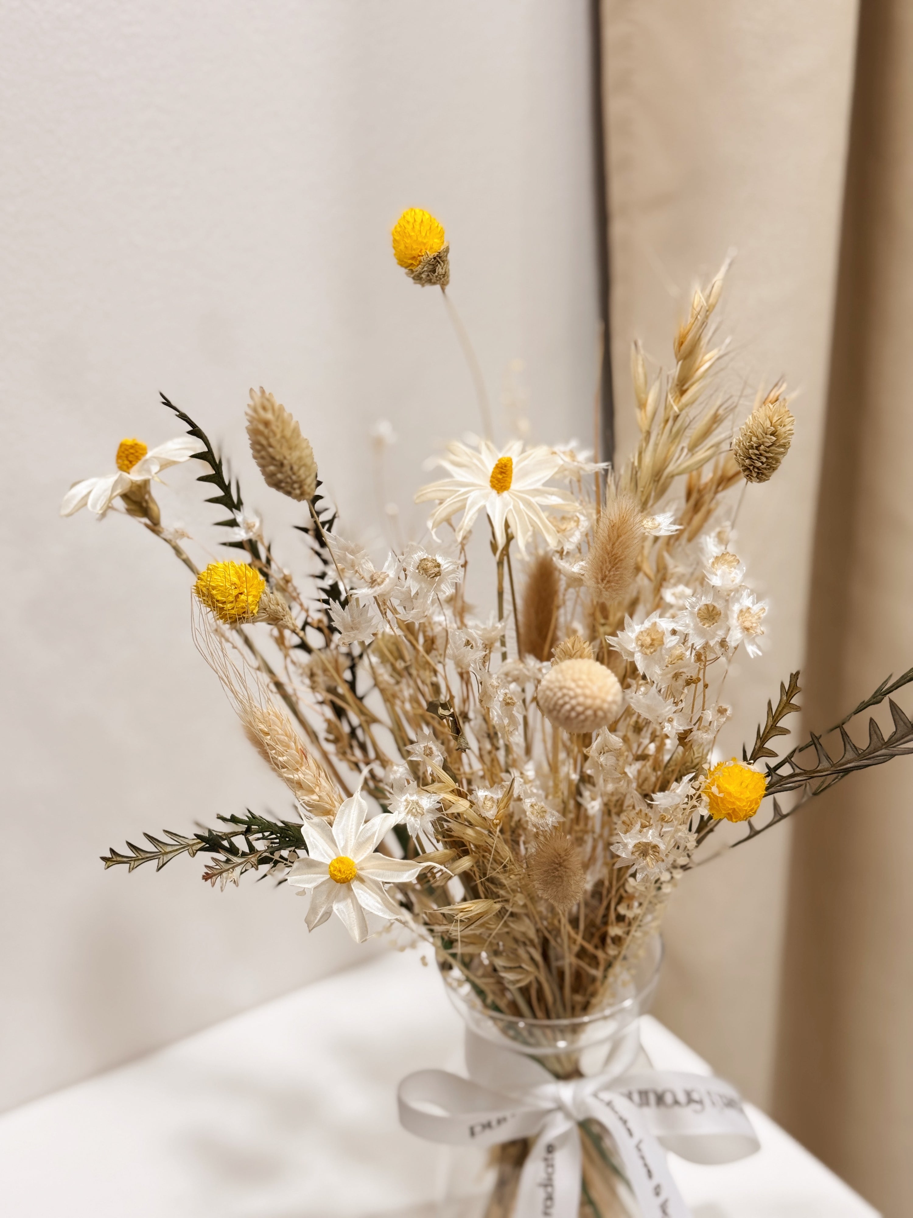 Vase arrangement of dried daisies with white and yellow filler flower, arranged in a clear glass vase, crafted by Eden's Ground.