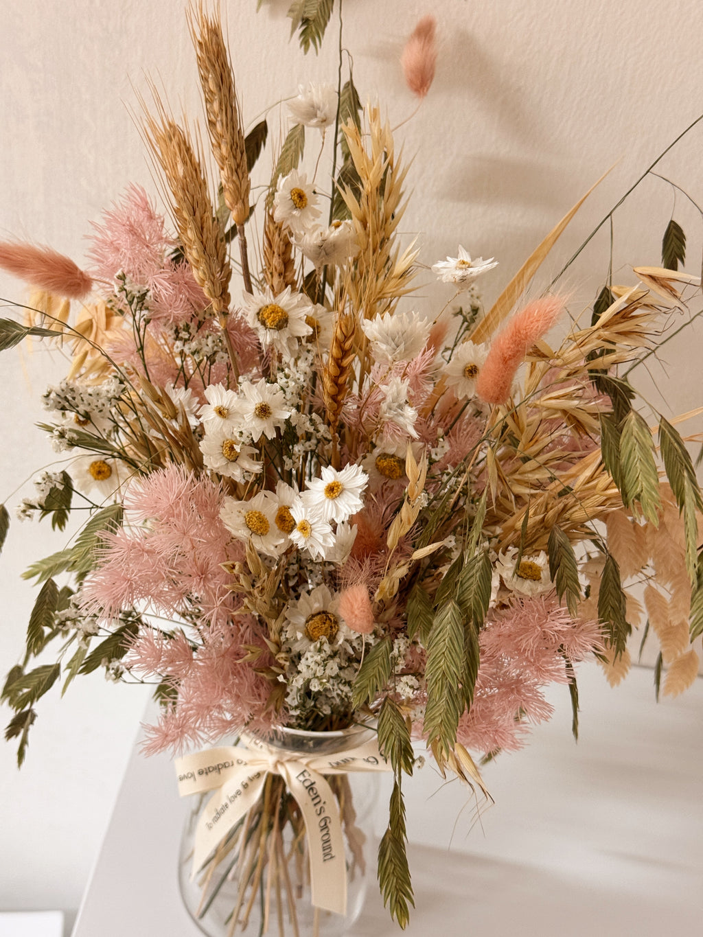 A preserved and dried flower arrangement in a clear glass jar with a cream ribbon, featuring pink asparagus fern, wheat stalks, daisies, and green foliage. Low-maintenance and long-lasting, it's perfect as a housewarming gift or as a timeless piece of home décor.