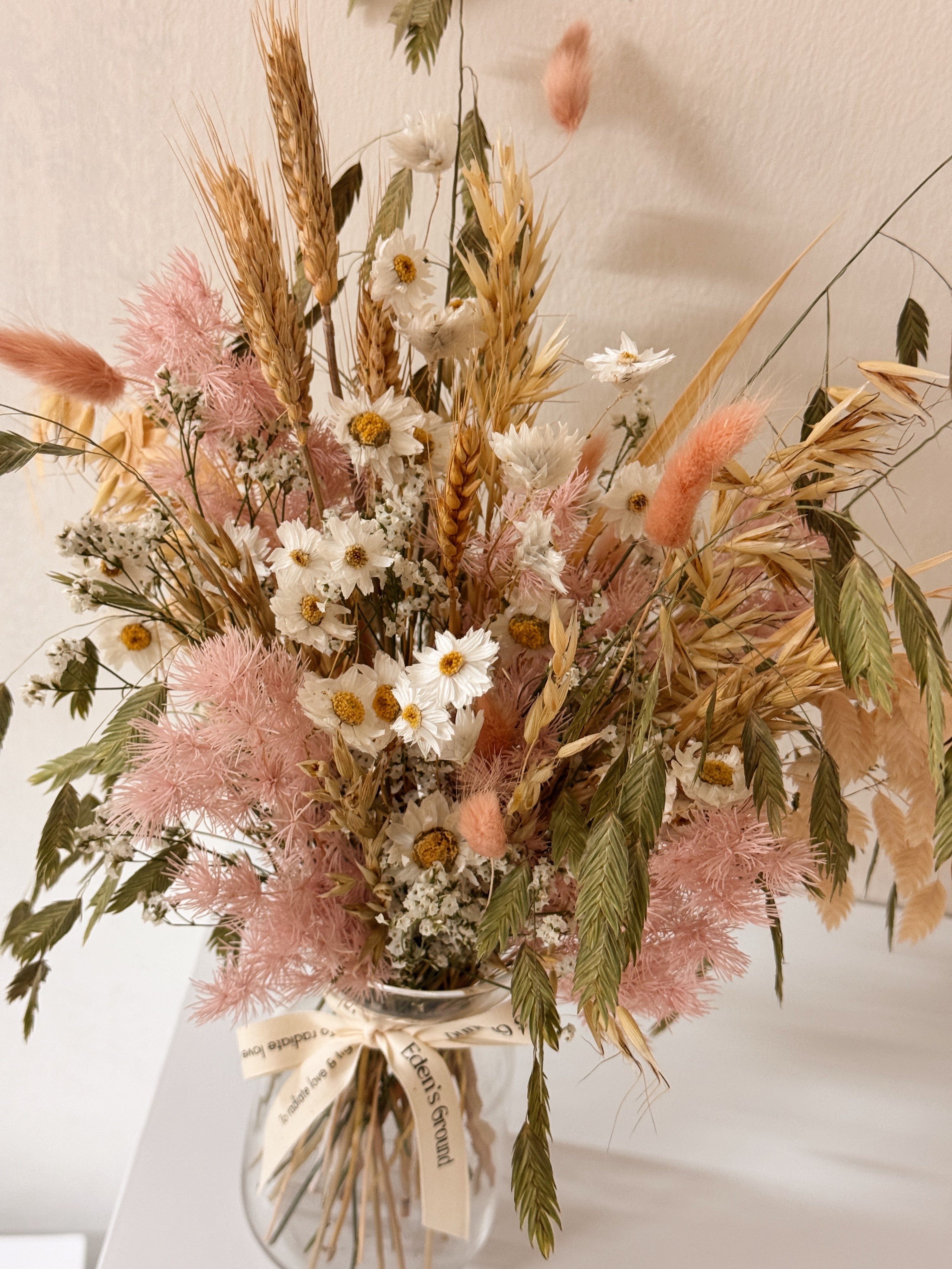 A preserved and dried flower arrangement in a clear glass jar with a cream ribbon, featuring pink asparagus fern, wheat stalks, daisies, and green foliage. Low-maintenance and long-lasting, it's perfect as a housewarming gift or as a timeless piece of home décor.