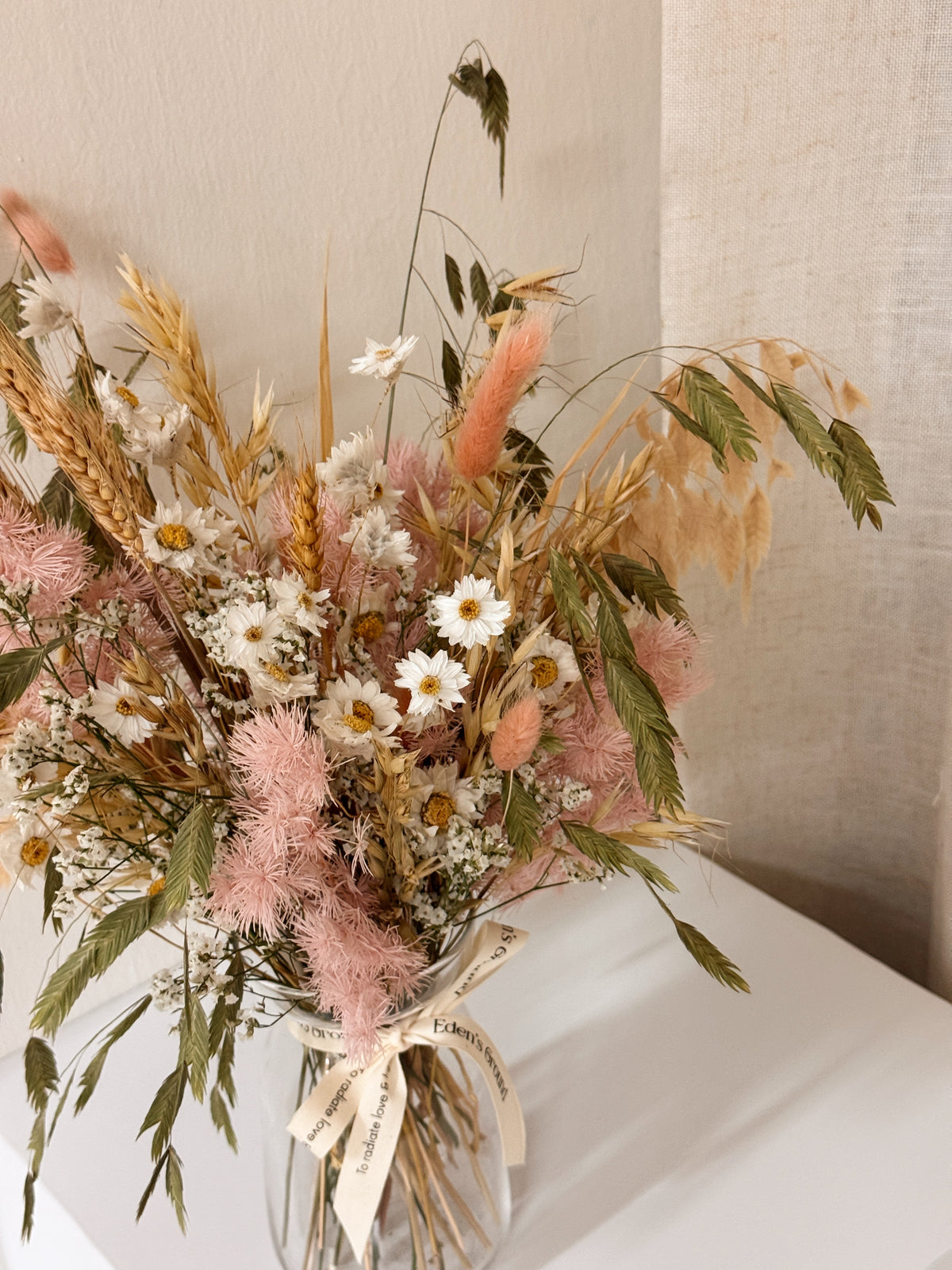 A preserved and dried flower arrangement in a clear glass jar with a cream ribbon, featuring pink asparagus fern, wheat stalks, daisies, and green foliage. Low-maintenance and long-lasting, it's perfect as a housewarming gift or as a timeless piece of home décor.