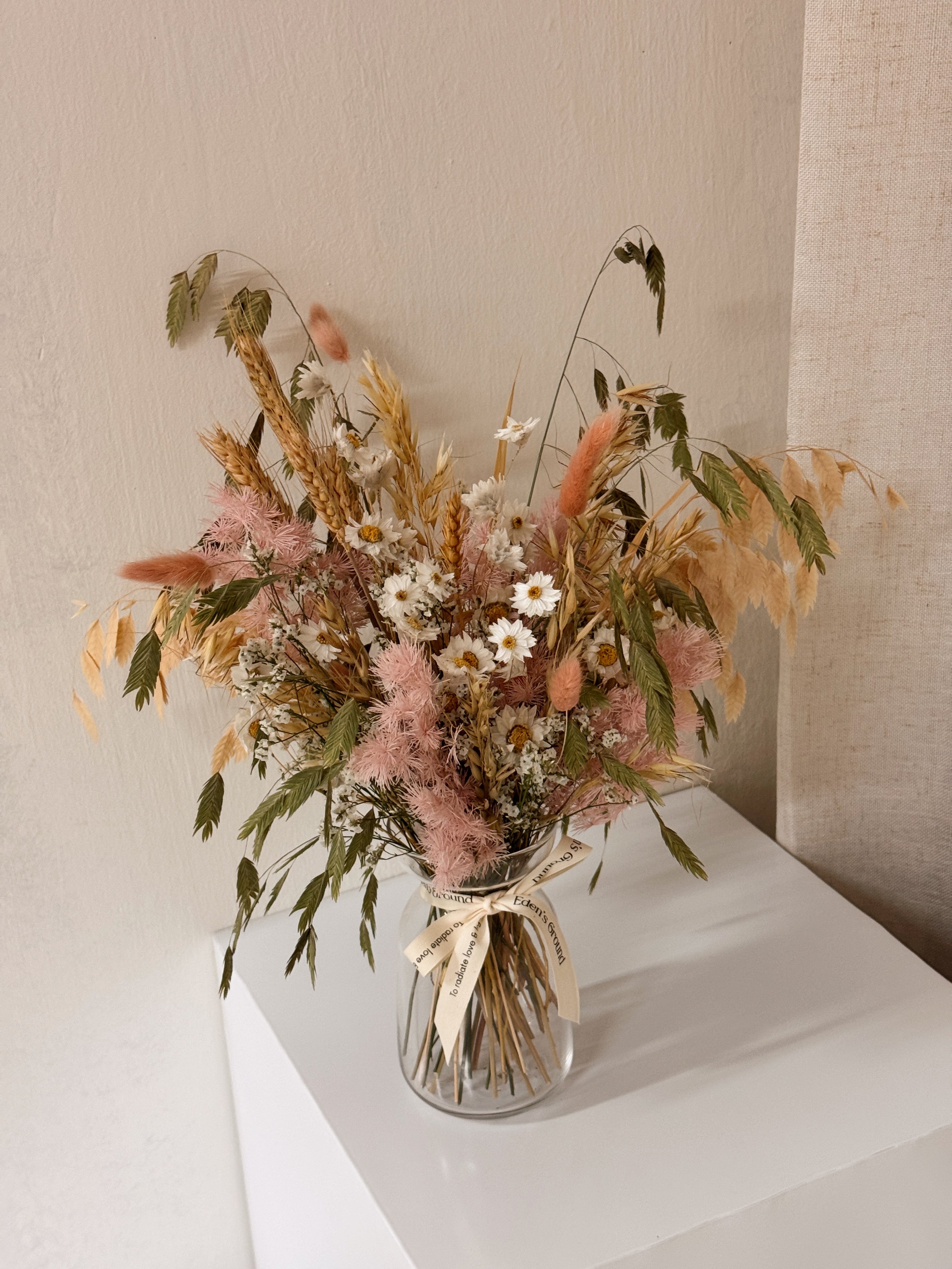A preserved and dried flower arrangement in a clear glass jar with a cream ribbon, featuring pink asparagus fern, wheat stalks, daisies, and green foliage. Low-maintenance and long-lasting, it's perfect as a housewarming gift or as a timeless piece of home décor.