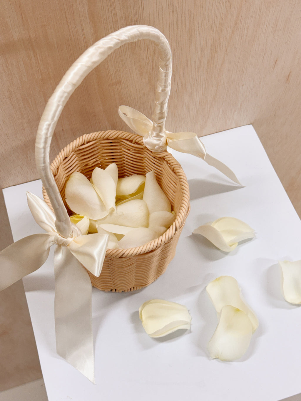 A small woven wicker flower basket with a white satin handle and bow, filled with white rose petals, ready for wedding march in for flower girl to toss the petals to welcome the groom and bride.