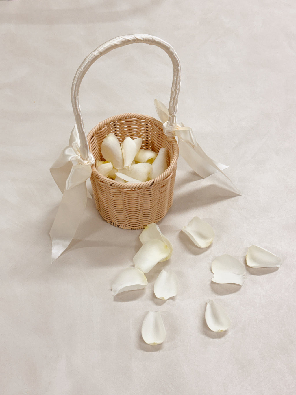 A small woven wicker flower basket with a white satin handle and bow, filled with white rose petals, ready for wedding march in for flower girl to toss the petals to welcome the groom and bride.