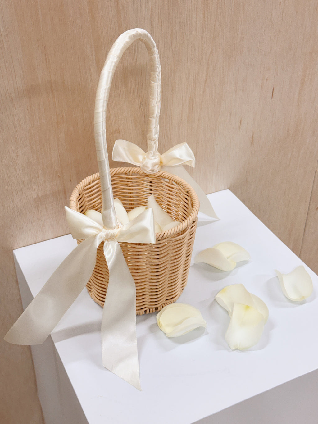 A small woven wicker flower basket with a white satin handle and bow, filled with white rose petals, ready for wedding march in for flower girl to toss the petals to welcome the groom and bride.
