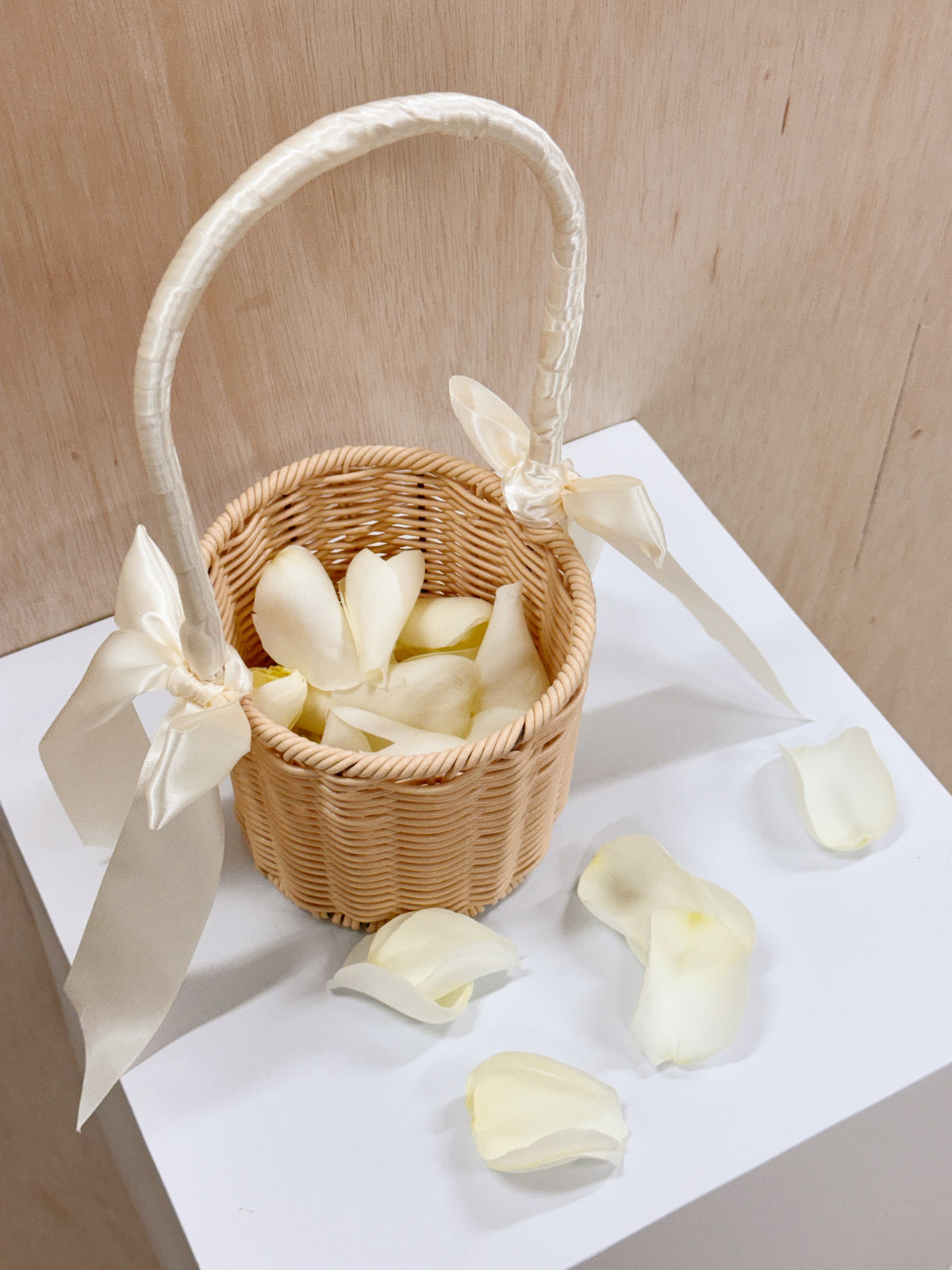 A small woven wicker flower basket with a white satin handle and bow, filled with white rose petals, ready for wedding march in for flower girl to toss the petals to welcome the groom and bride.