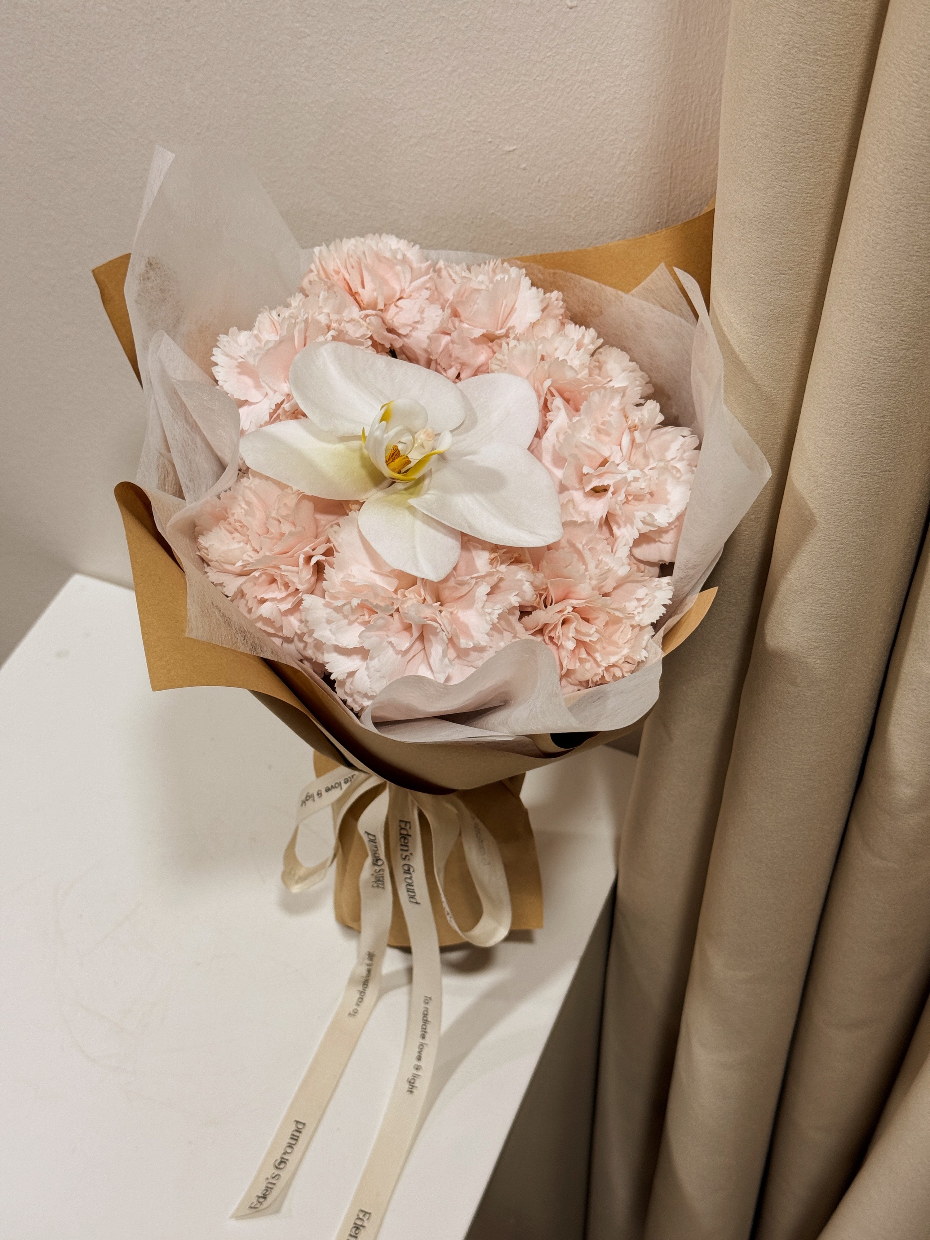 Gentle Touch of Grace bouquet, for Mother's Day, made with fresh sweet pink Carnations along with a white Phalaenopsis Orchid, wrapped in white tissue and brown kraft paper crafted by Eden’s Ground, a floral design boutique based in Singapore.