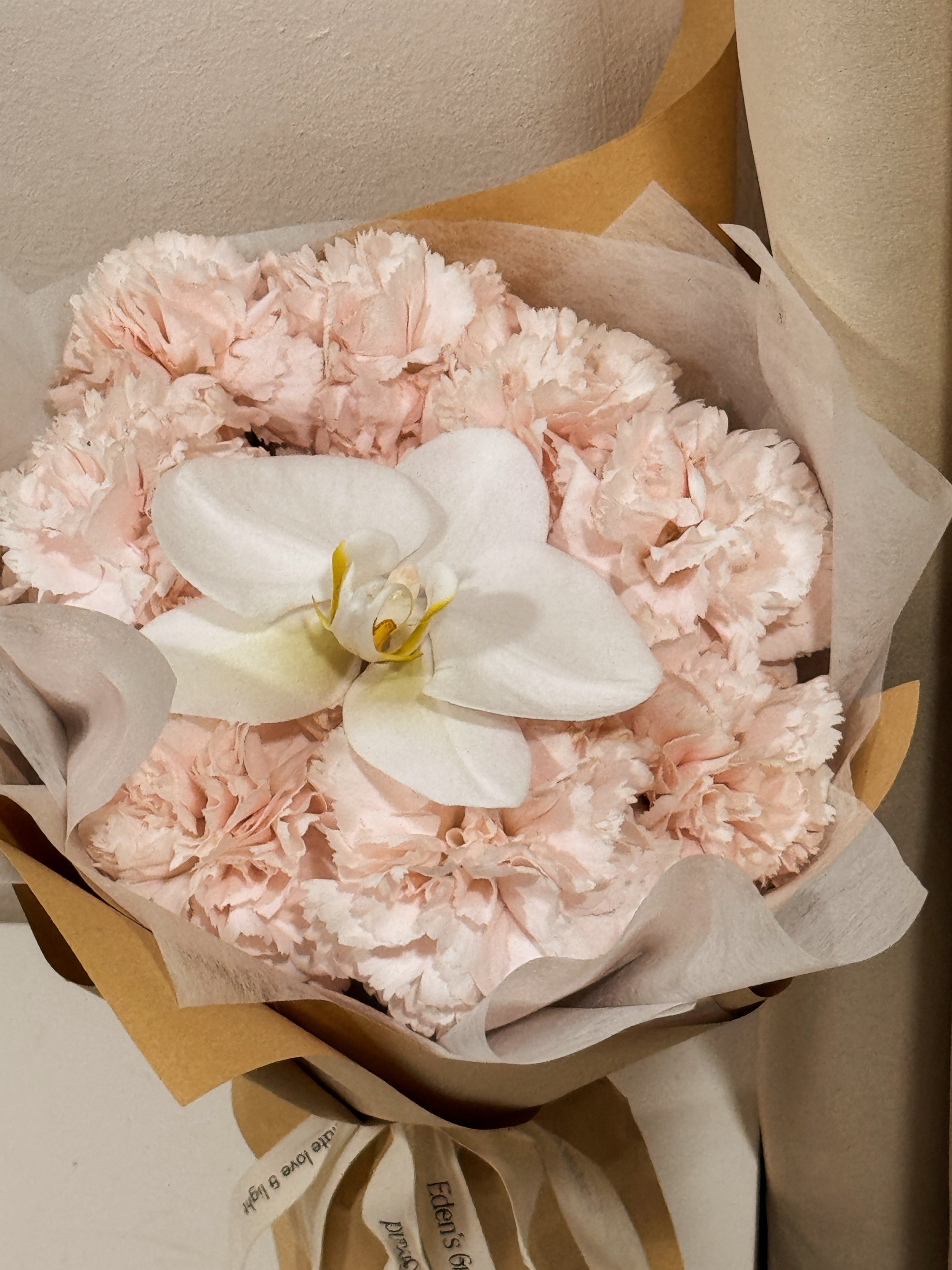 Gentle Touch of Grace bouquet, for Mother's Day, made with fresh sweet pink Carnations along with a white Phalaenopsis Orchid, wrapped in white tissue and brown kraft paper crafted by Eden’s Ground, a floral design boutique based in Singapore.