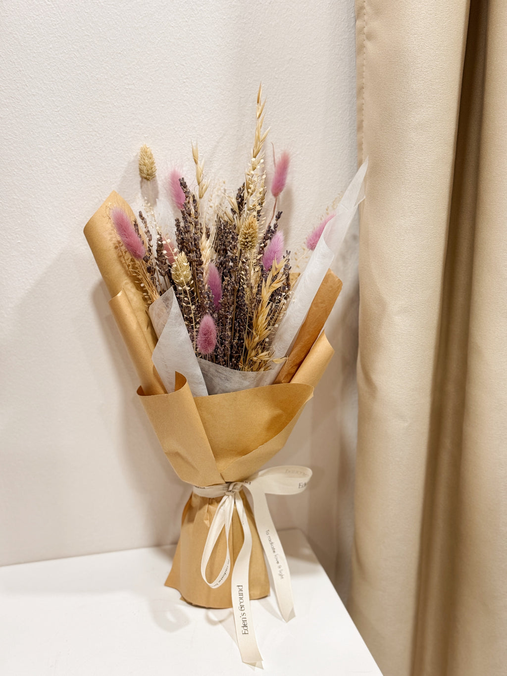 Lavender Bunch is a delicate bouquet arrangement of dried lavender flowers, gathered in a clear glass vase and tied with a white ribbon, crafted by Eden’s Ground, a floral design boutique.