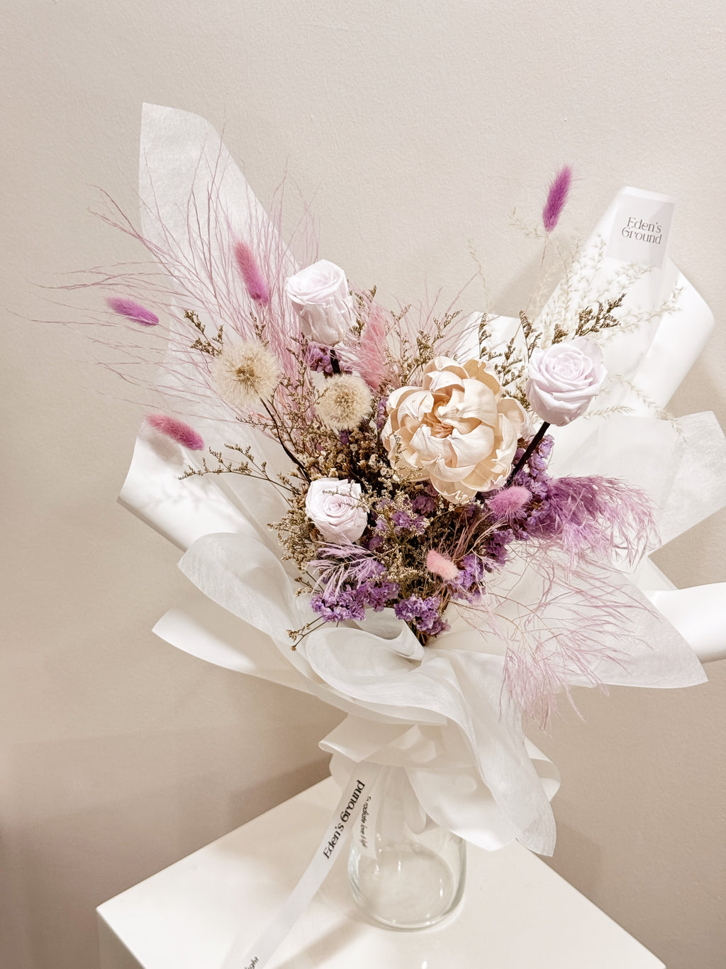 Lavender Haze bouquet made with preserved roses along with dandelions, filler flowers and foliage, wrapped in white tissue and white glassine paper crafted by Eden’s Ground.