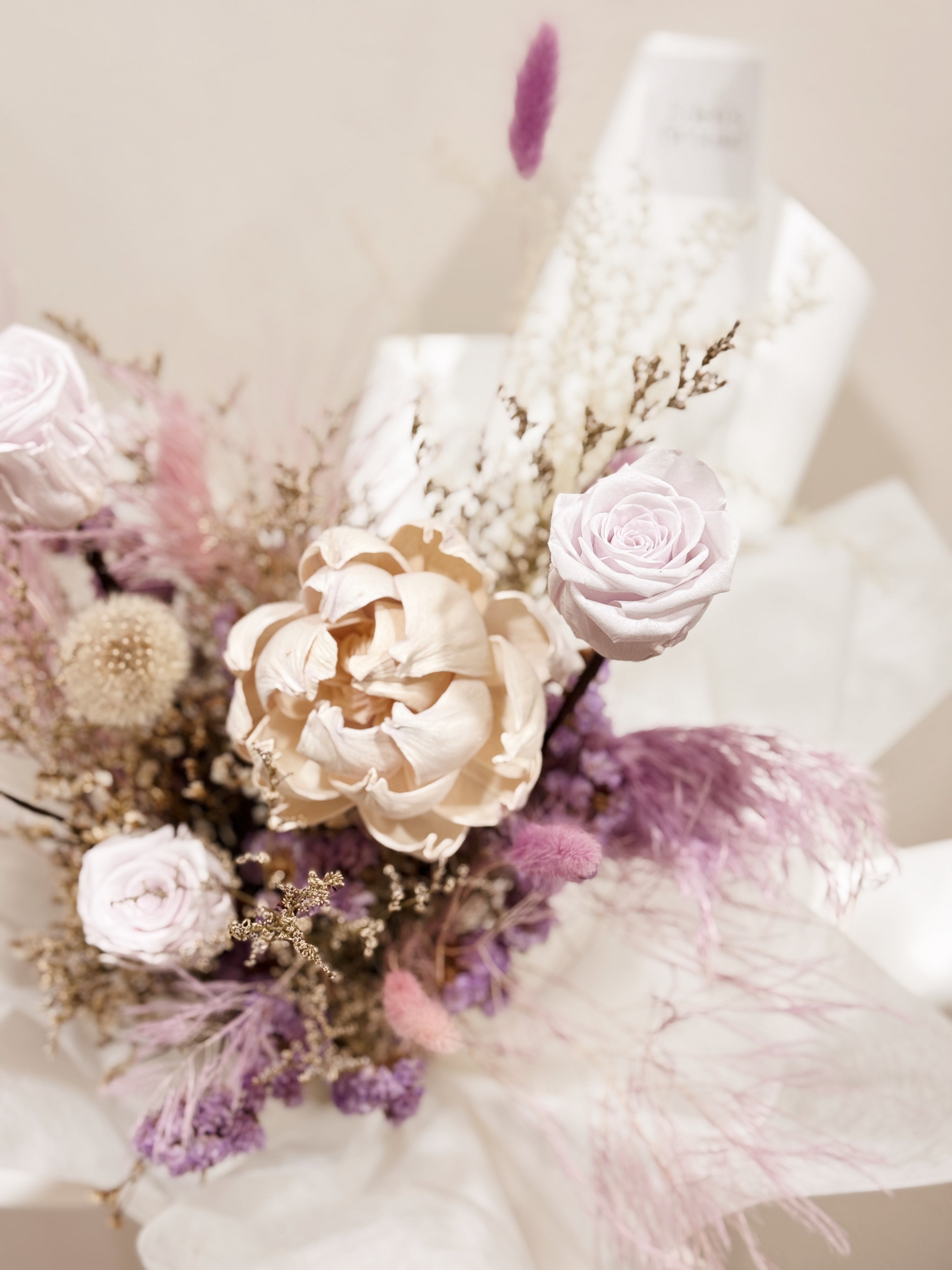 Lavender Haze bouquet made with preserved roses along with dandelions, filler flowers and foliage, wrapped in white tissue and white glassine paper crafted by Eden’s Ground.