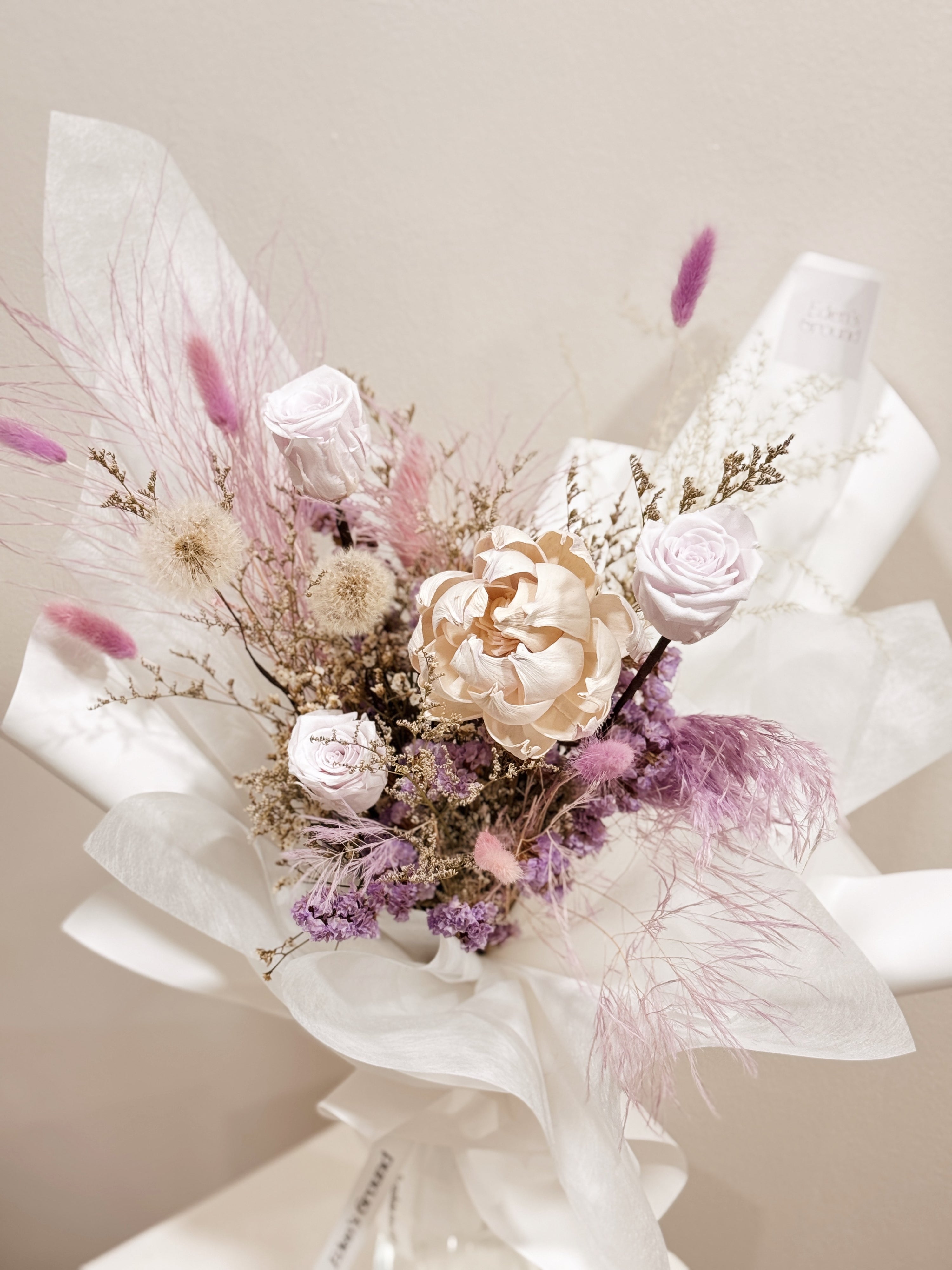 Lavender Haze bouquet made with preserved roses along with dandelions, filler flowers and foliage, wrapped in white tissue and white glassine paper crafted by Eden’s Ground.