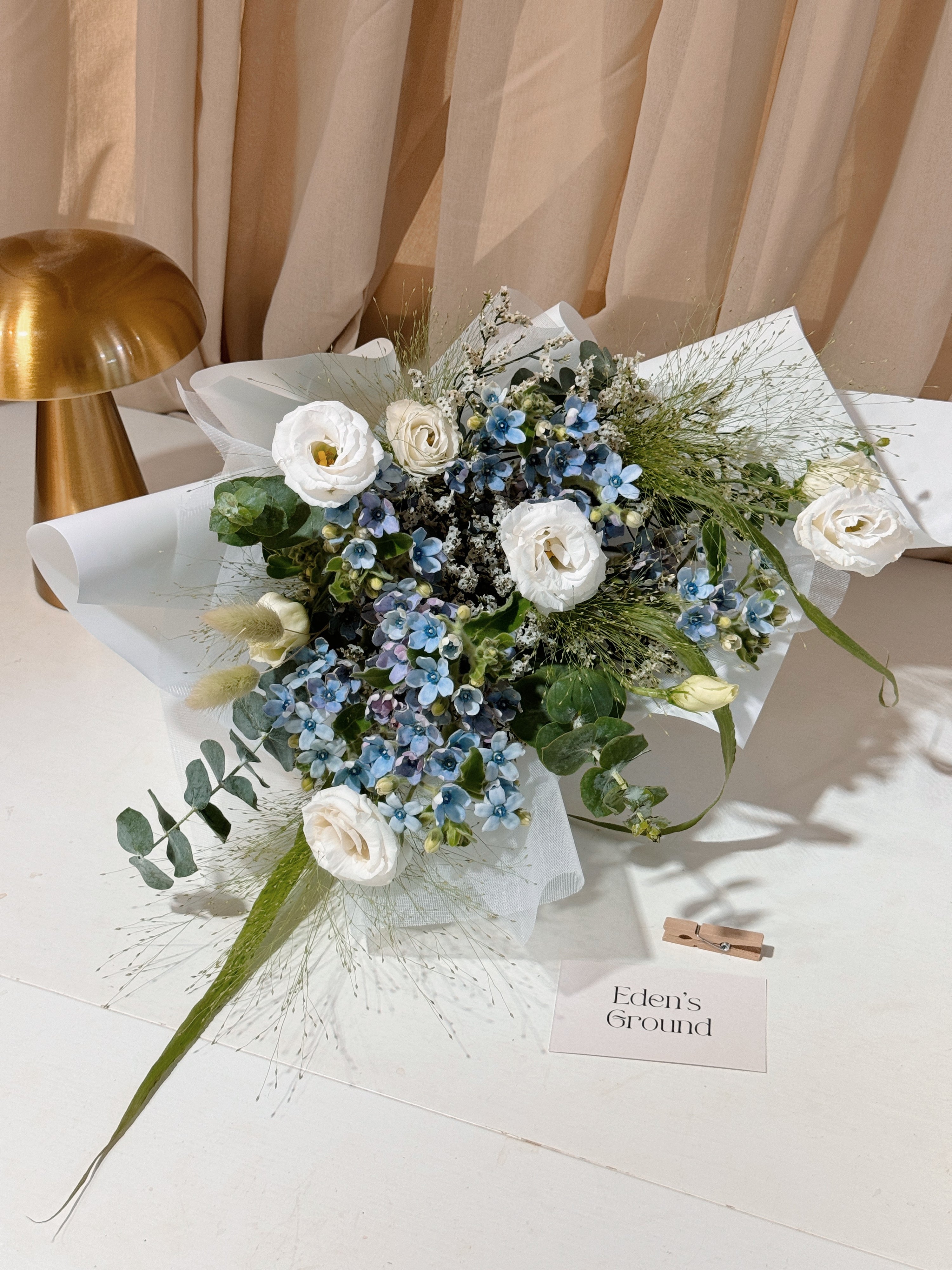 My Daystar blue tweedia bouquet featuring white eustoma, panicum and eucalyptus foliage, wrapped in white tissue paper and white glassine paper crafted by Eden’s Ground.