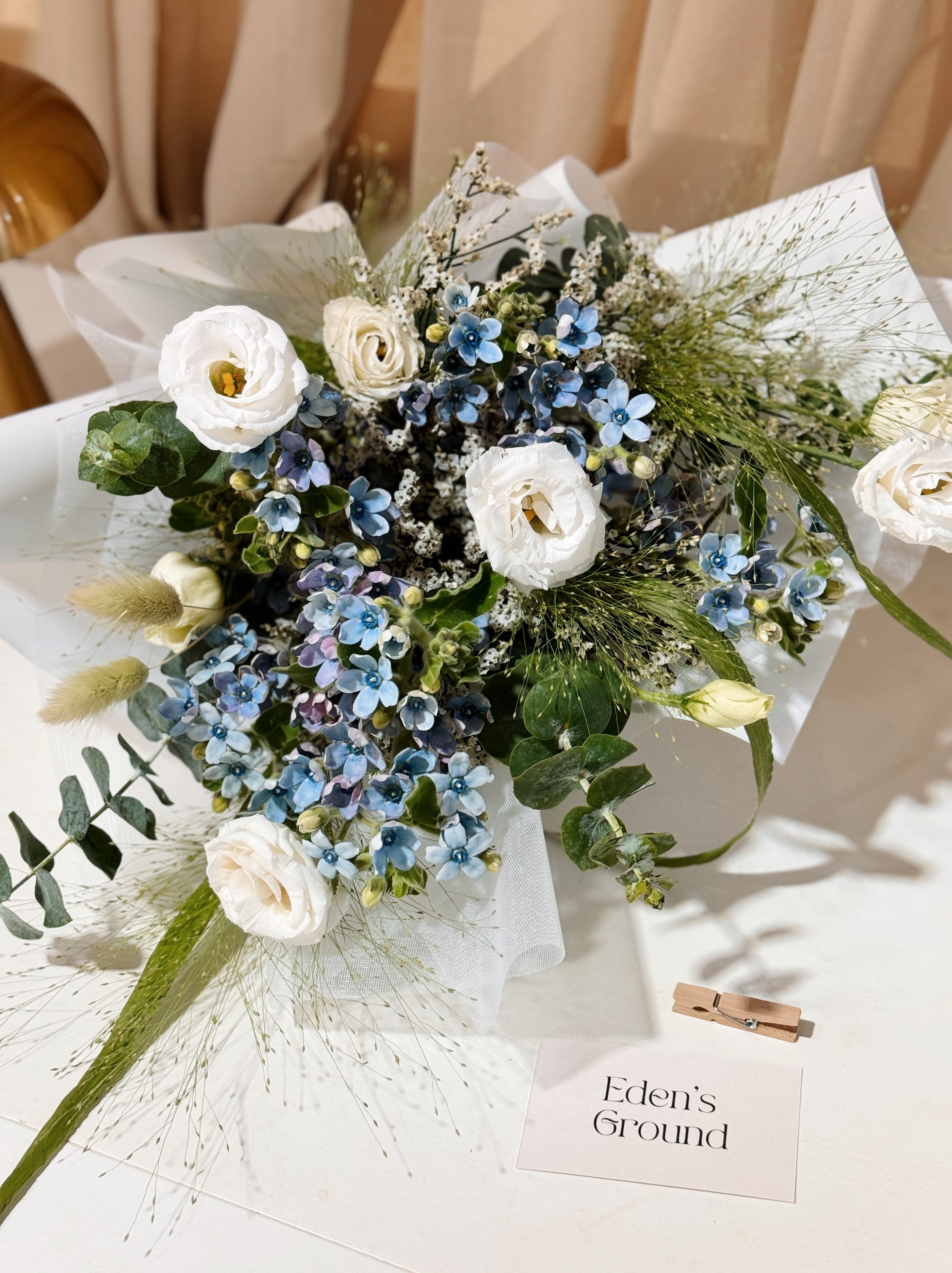 My Daystar blue tweedia bouquet featuring white eustoma, panicum and eucalyptus foliage, wrapped in white tissue paper and white glassine paper crafted by Eden’s Ground.