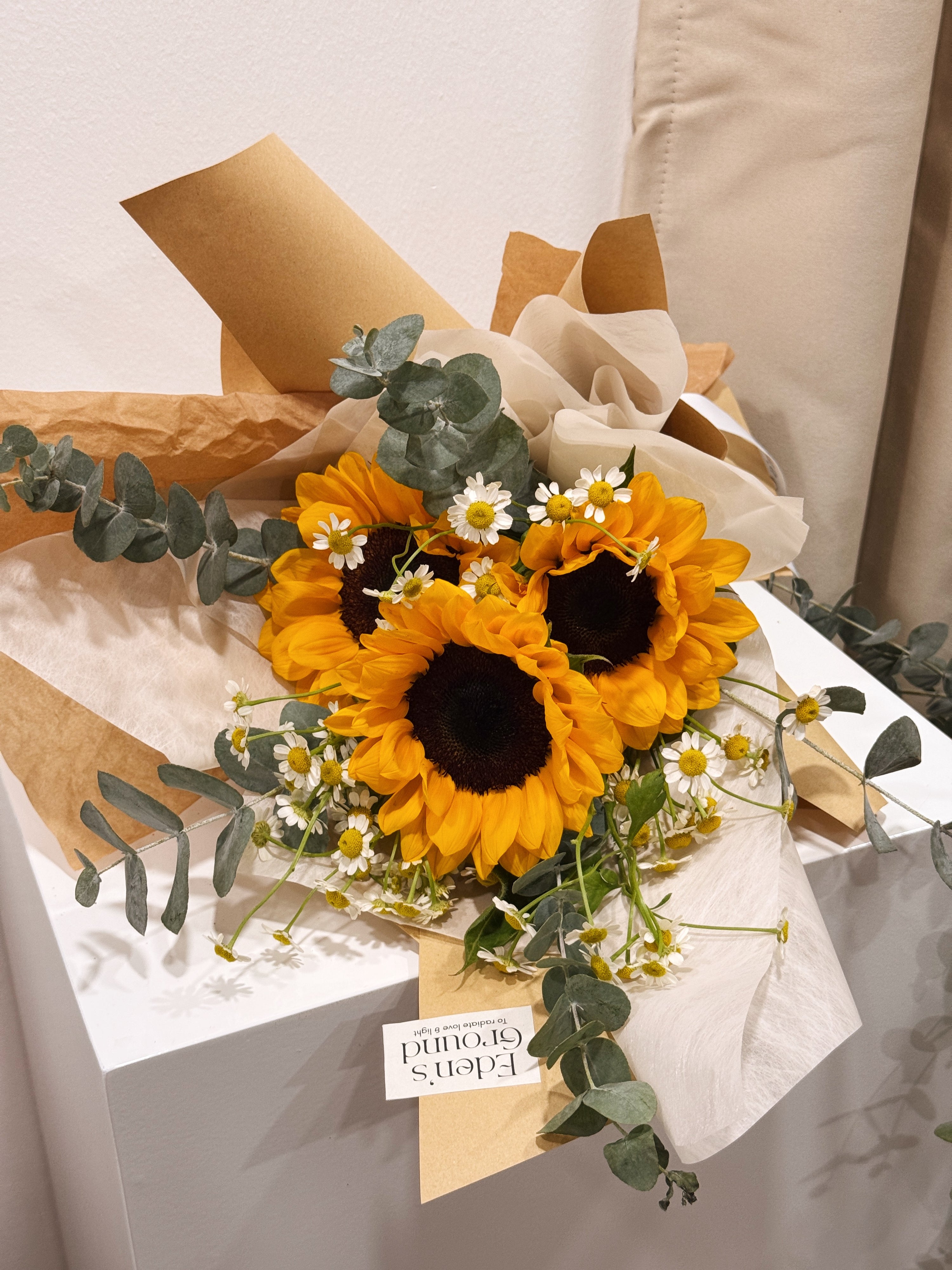 Sunflower bouquet featuring chamomile daisies and eucalyptus foliage, wrapped in tissue paper and brown kraft paper crafted by Eden’s Ground.