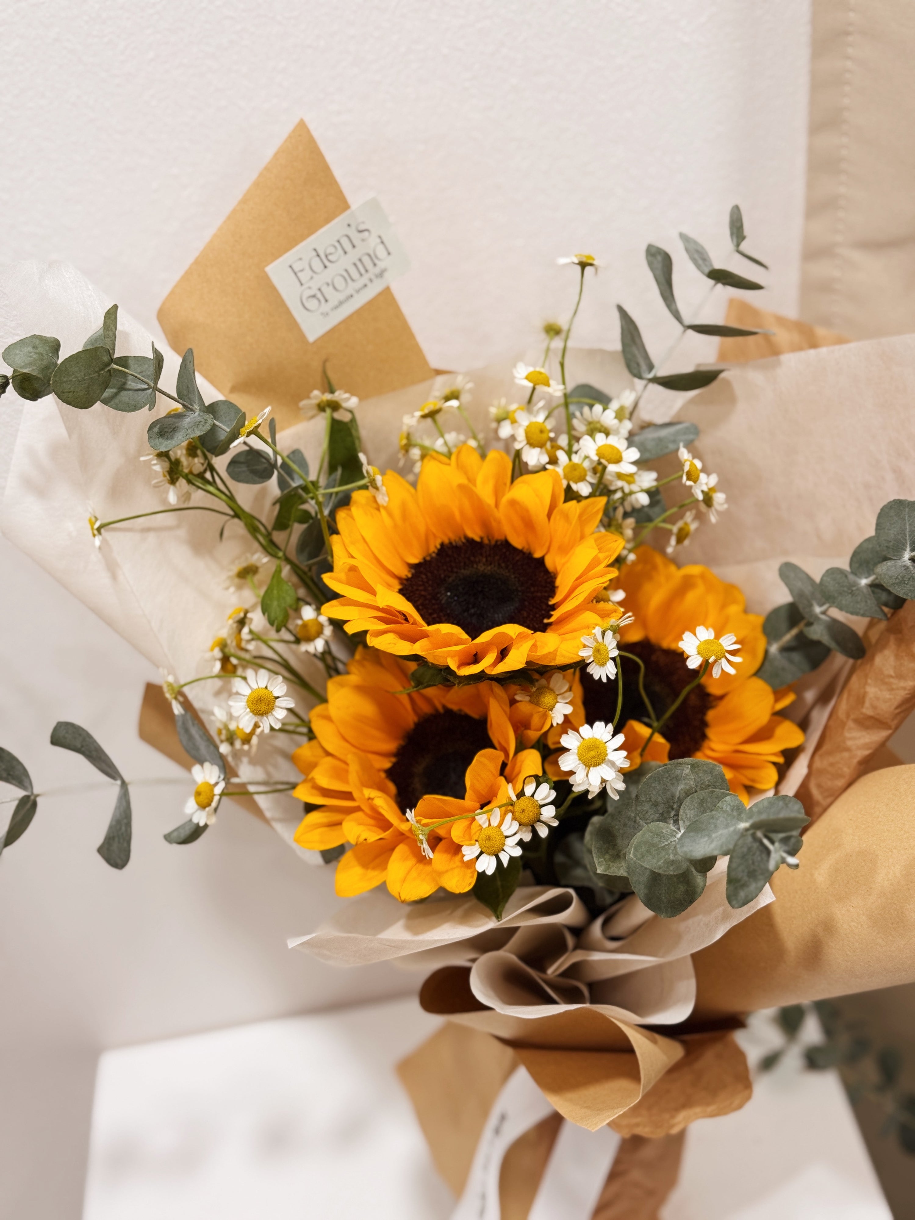 Sunflower bouquet featuring chamomile daisies and eucalyptus foliage, wrapped in tissue paper and brown kraft paper crafted by Eden’s Ground.