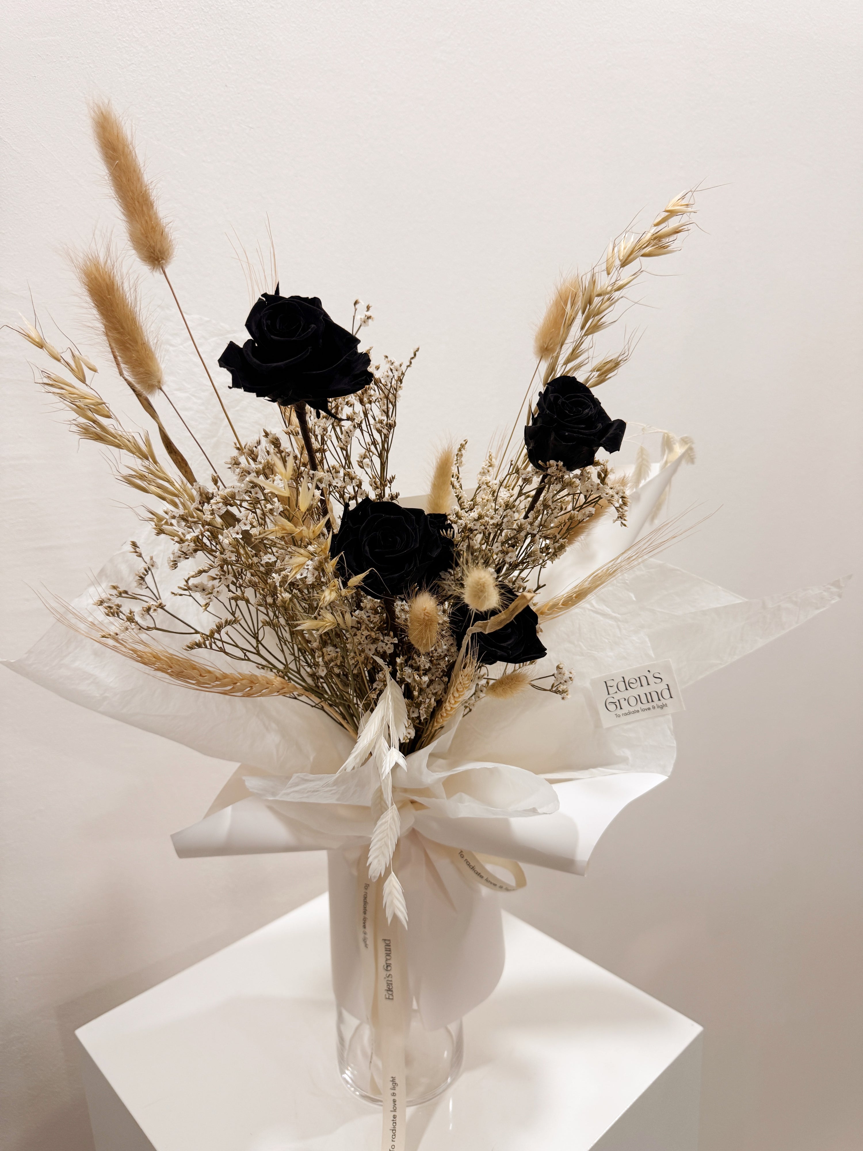 Noir Muse bouquet made with preserved black roses along with filler flowers and foliage, wrapped in white paper crafted by Eden’s Ground, a floral design boutique.
