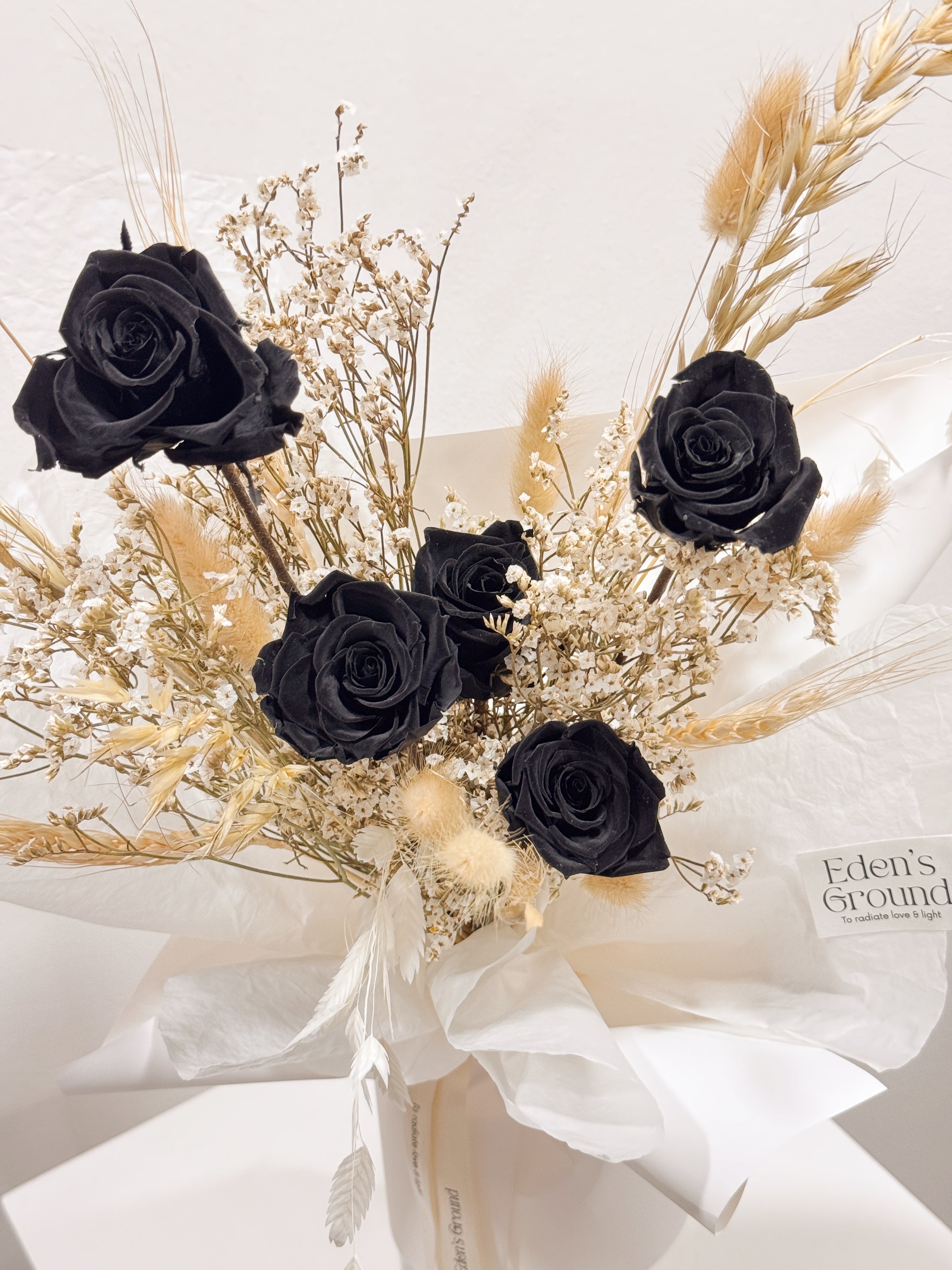 Noir Muse bouquet made with preserved black roses along with filler flowers and foliage, wrapped in white paper crafted by Eden’s Ground, a floral design boutique.