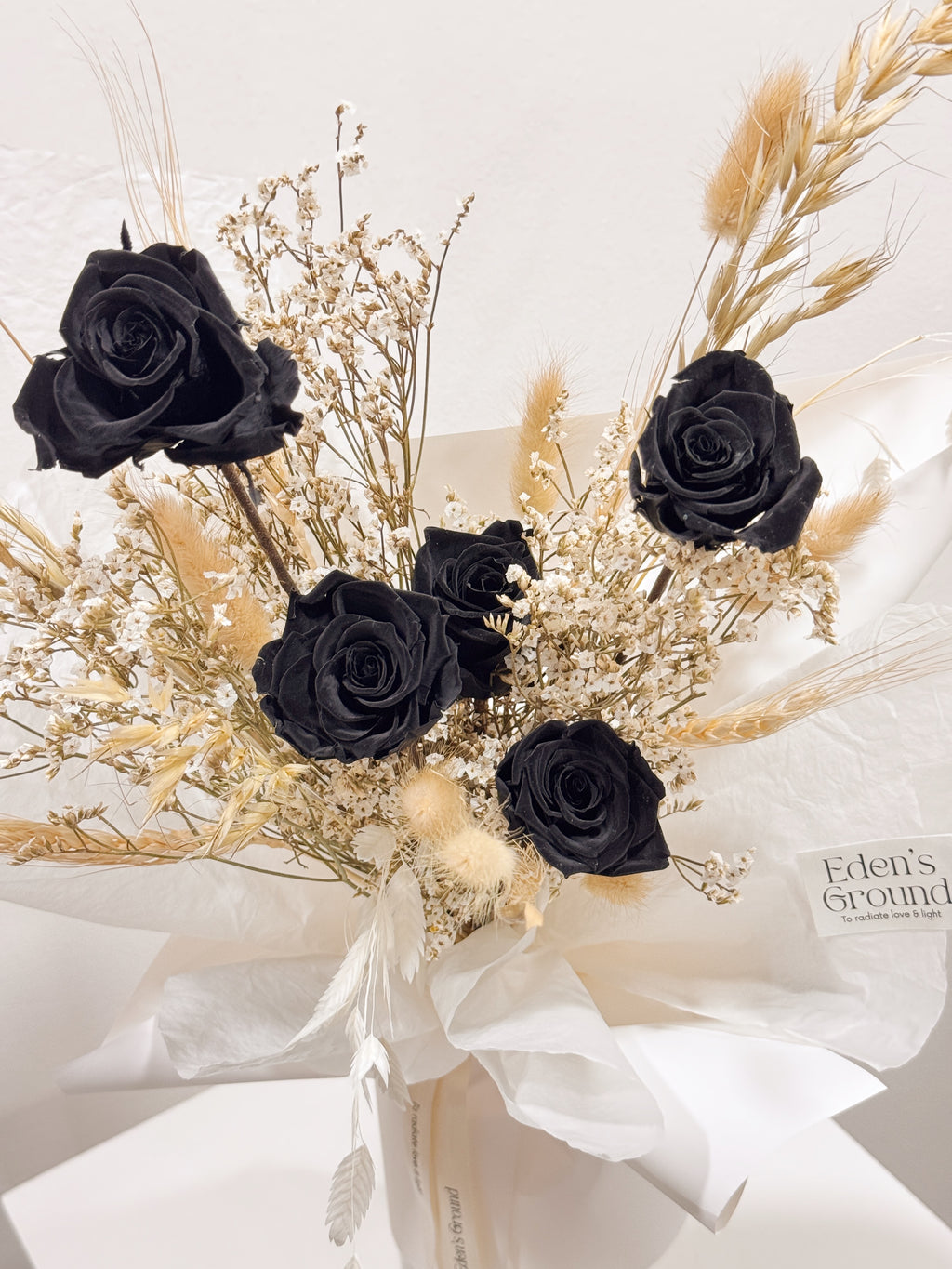 Noir Muse bouquet made with preserved black roses along with filler flowers and foliage, wrapped in white paper crafted by Eden’s Ground, a floral design boutique.