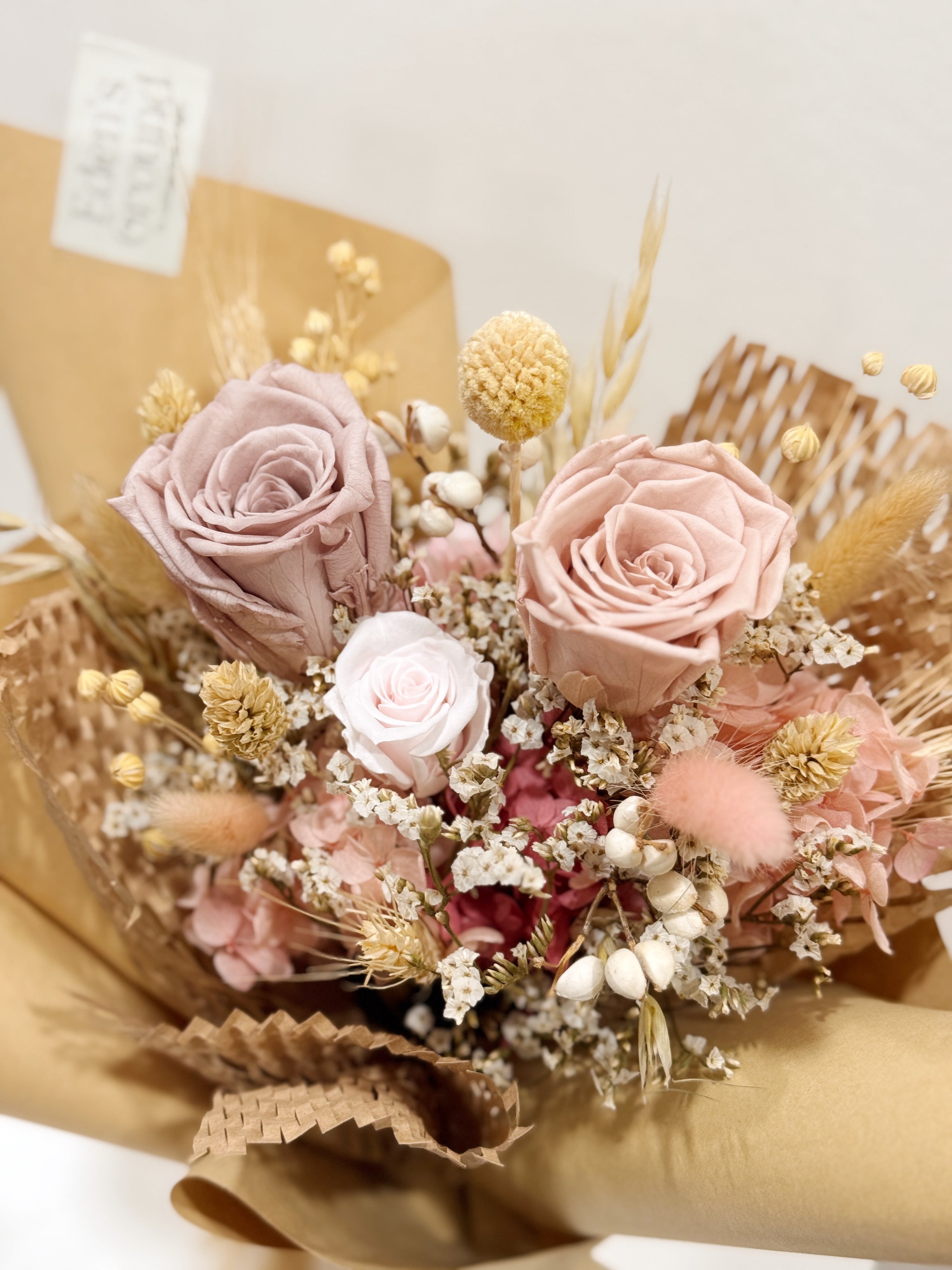 Petal Kiss bouquet made with preserved roses, hydranges along with filler flowers and foliage, wrapped in brown kraft paper crafted by Eden’s Ground, a floral design boutique.