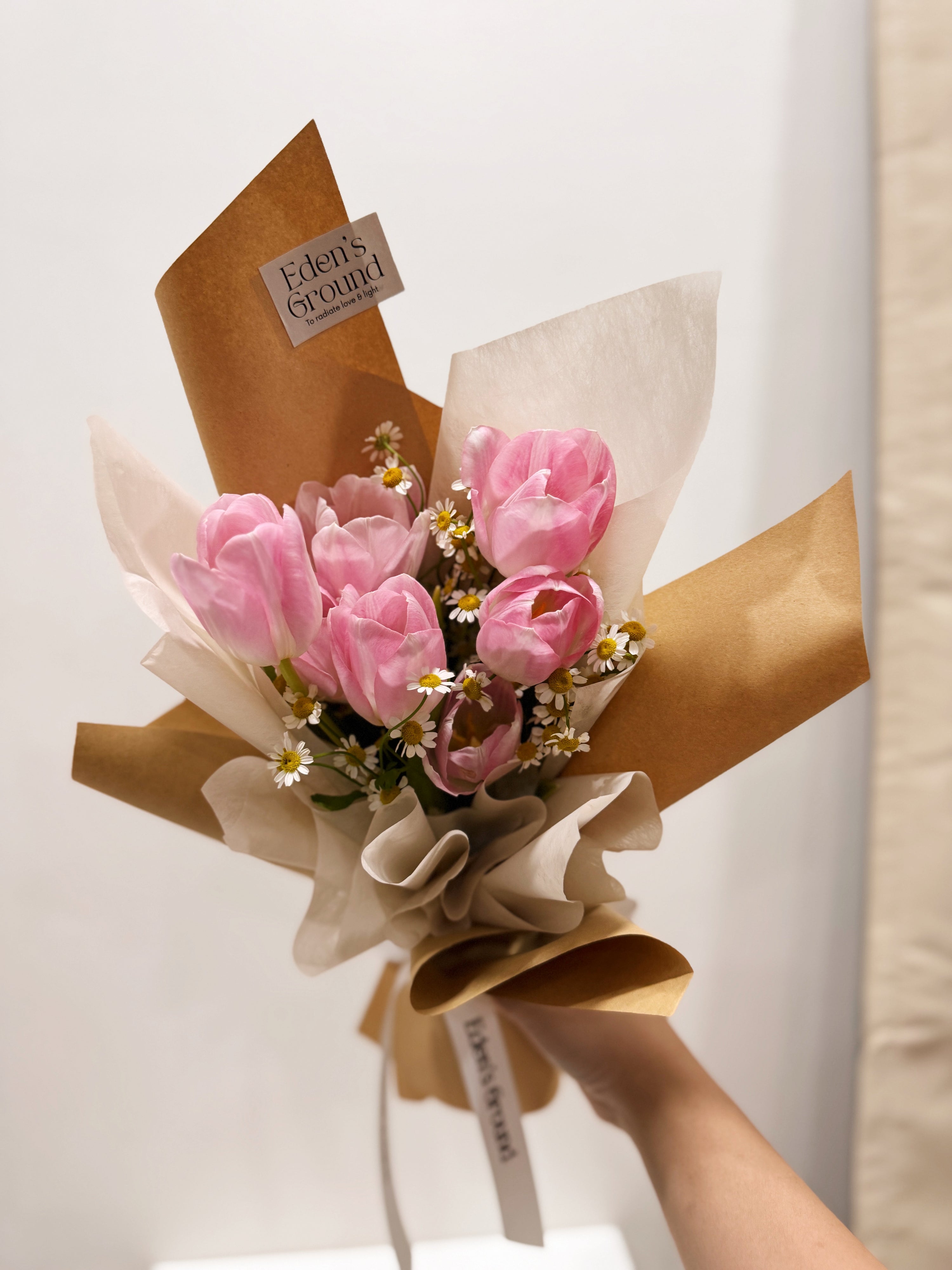 Pink tulip bouquet with chamomile daisies wrapped in tissue paper and brown kraft paper crafted by Eden’s Ground.