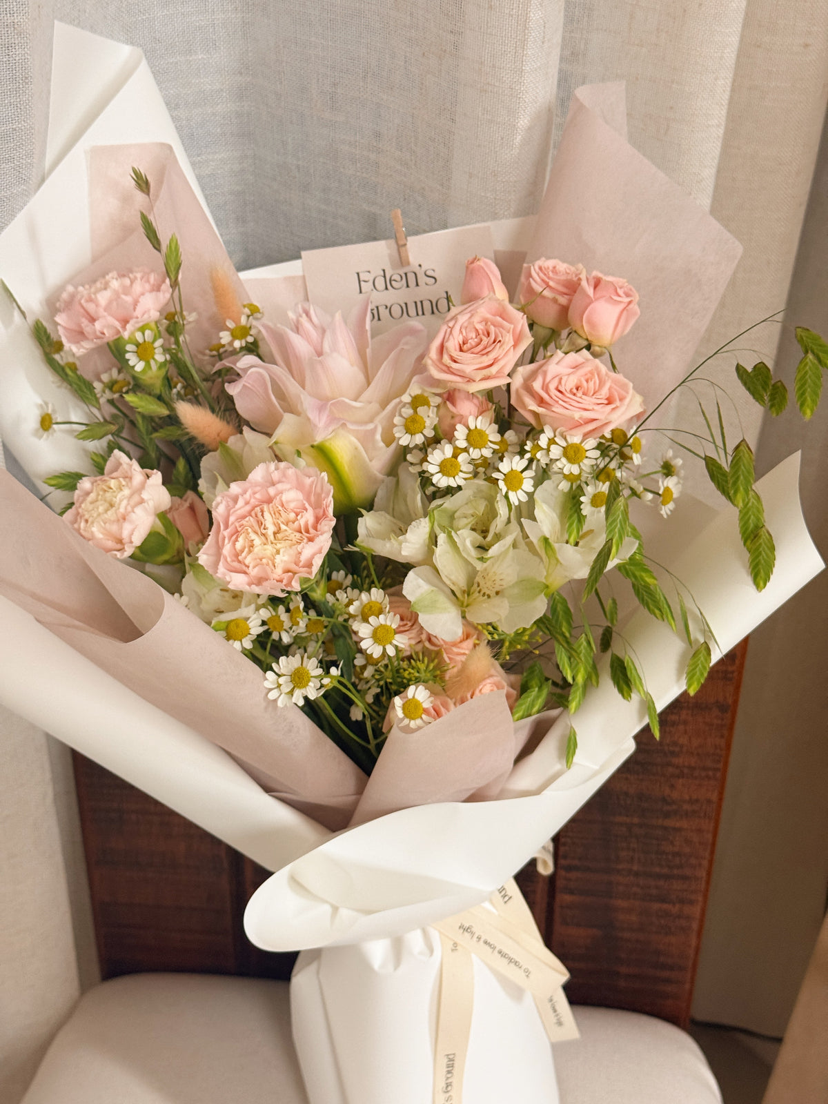 Poise Roselily featuring fragrant rose lily, carnation, roses, chamomile daisies and more with lots of textural foliage, wrapped in pink tissue paper and white glassine paper crafted by Eden’s Ground.