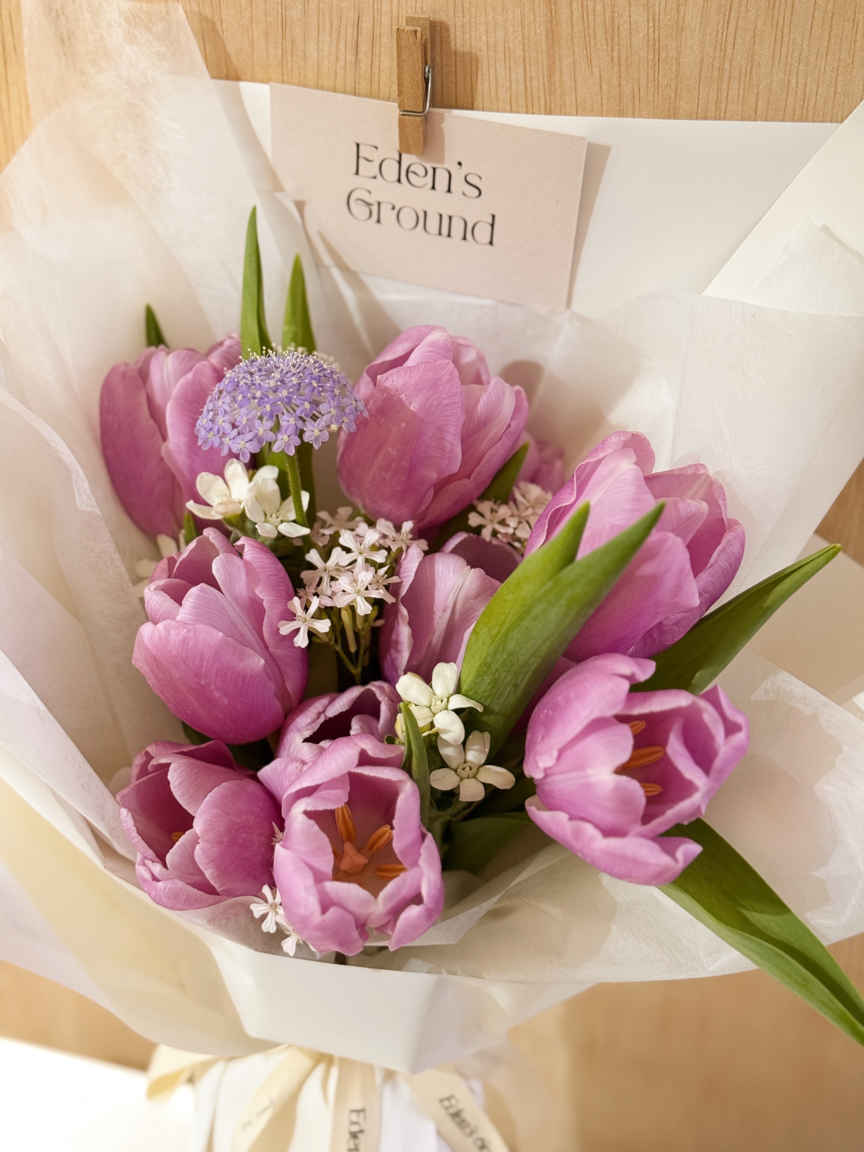 Purple tulip bouquet with filler flowers wrapped in tissue paper and white wrapper paper crafted by Eden’s Ground.