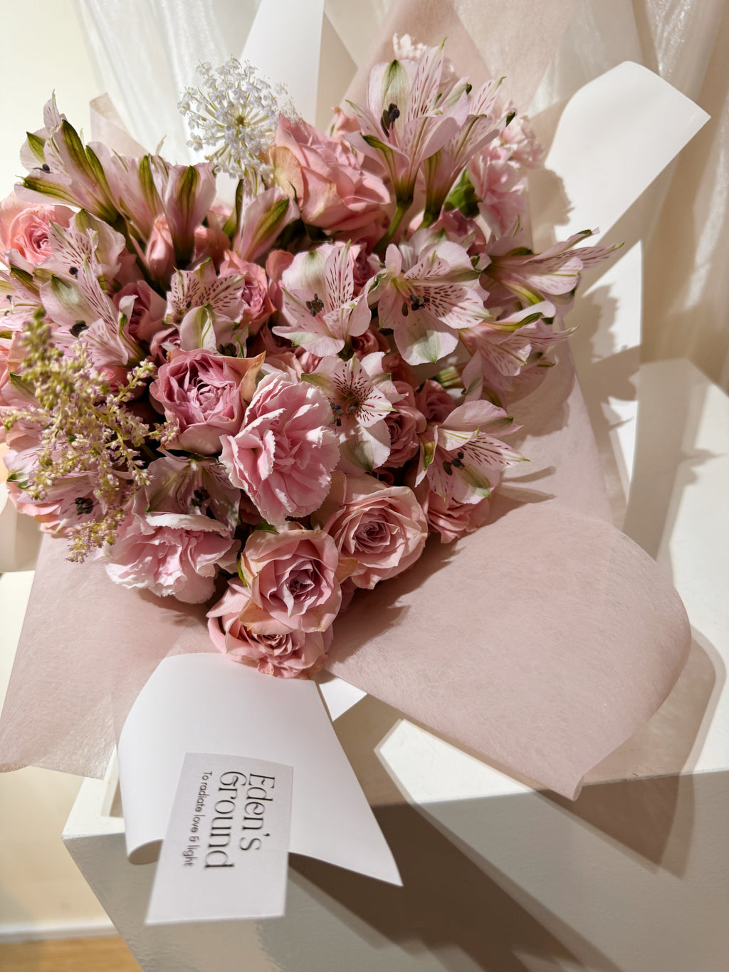 Rosy Devotion featuring sweet pink roses, alstroemeria lilies and carnation, wrapped in tissue paper and white glassing paper crafted by Eden’s Ground.
