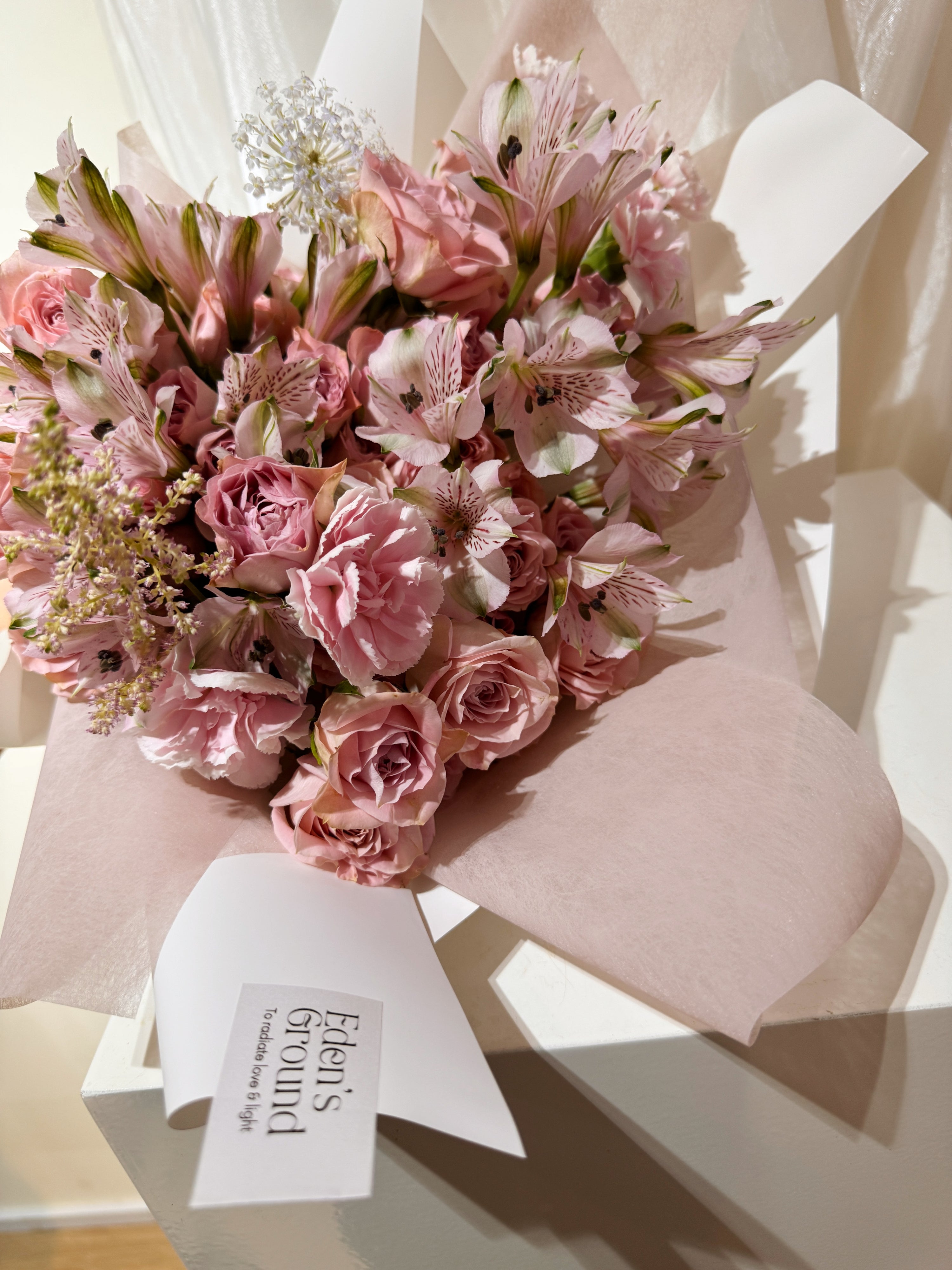 Rosy Devotion featuring sweet pink roses, alstroemeria lilies and carnation, wrapped in tissue paper and white glassing paper crafted by Eden’s Ground.