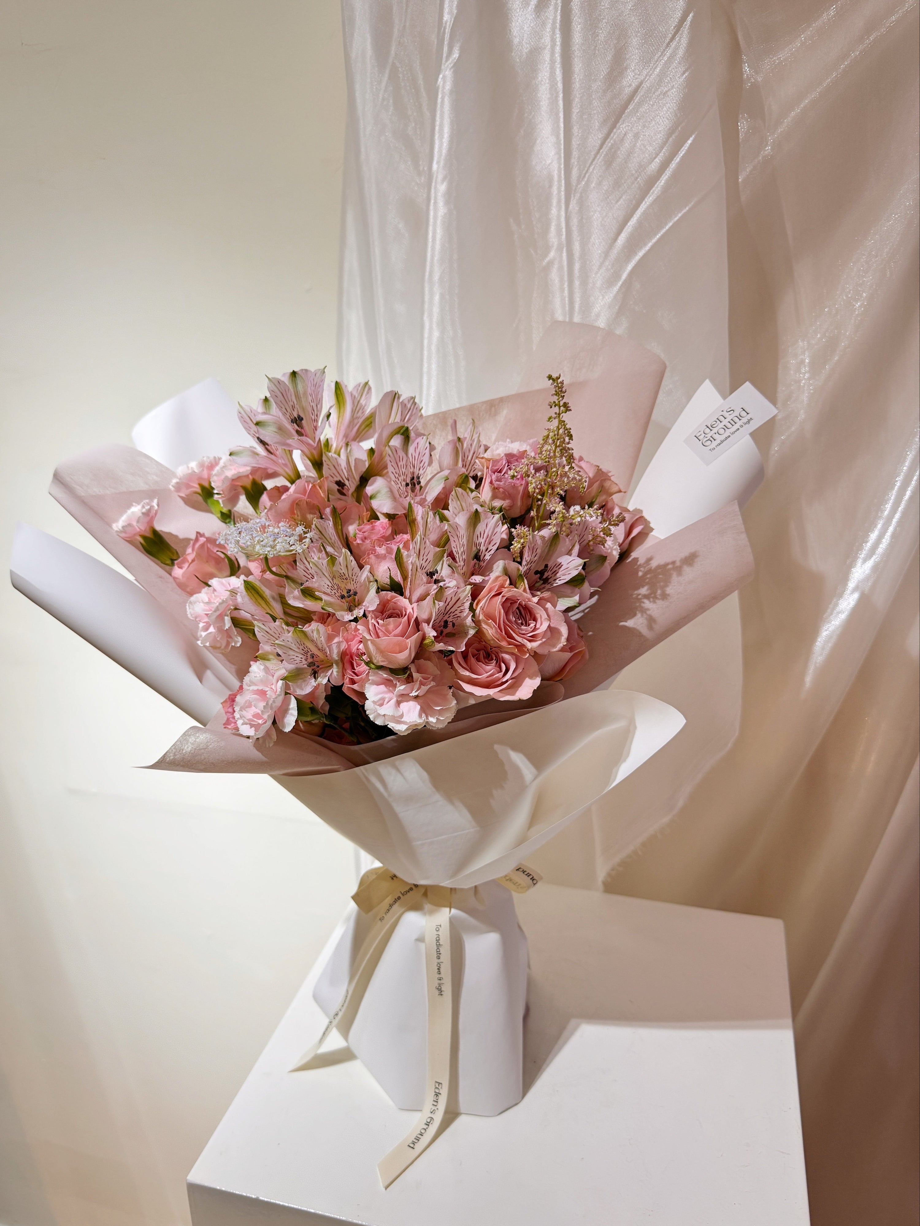 Rosy Devotion featuring sweet pink roses, alstroemeria lilies and carnation, wrapped in tissue paper and white glassing paper crafted by Eden’s Ground.
