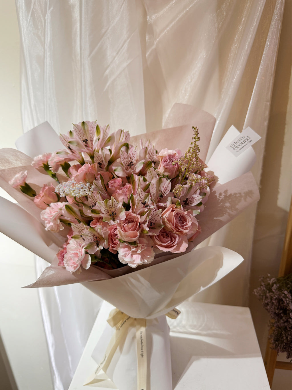 Rosy Devotion featuring sweet pink roses, alstroemeria lilies and carnation, wrapped in tissue paper and white glassing paper crafted by Eden’s Ground.