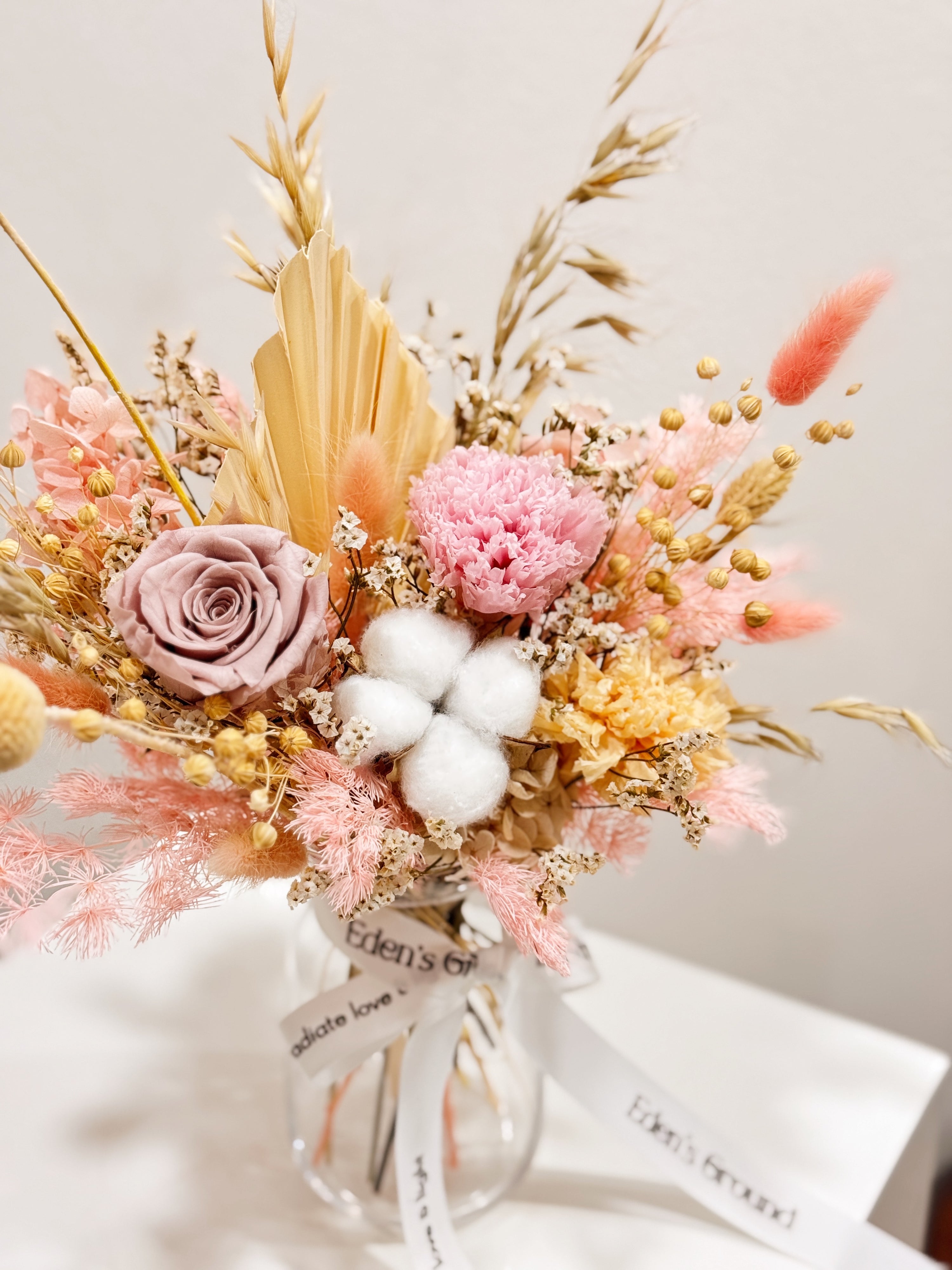 Soft Radiance, a preserved flower vase arrangement, featuring roses, carnations, fluffy cotton, and textural elements like bunny tails, grains, and palm spears, presented in a clear glass vase, crafted by Eden's Ground.
