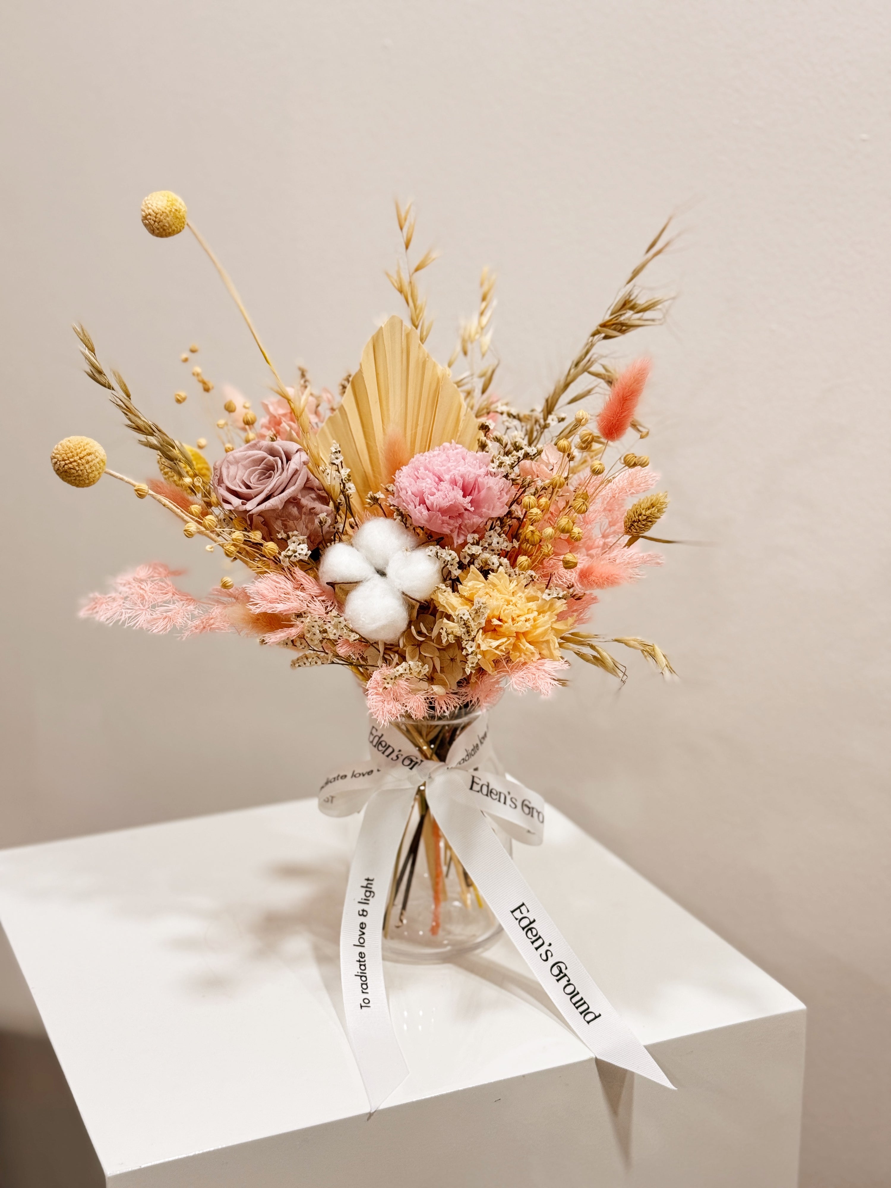 Soft Radiance, a preserved flower vase arrangement, featuring roses, carnations, fluffy cotton, and textural elements like bunny tails, grains, and palm spears, presented in a clear glass vase, crafted by Eden's Ground.