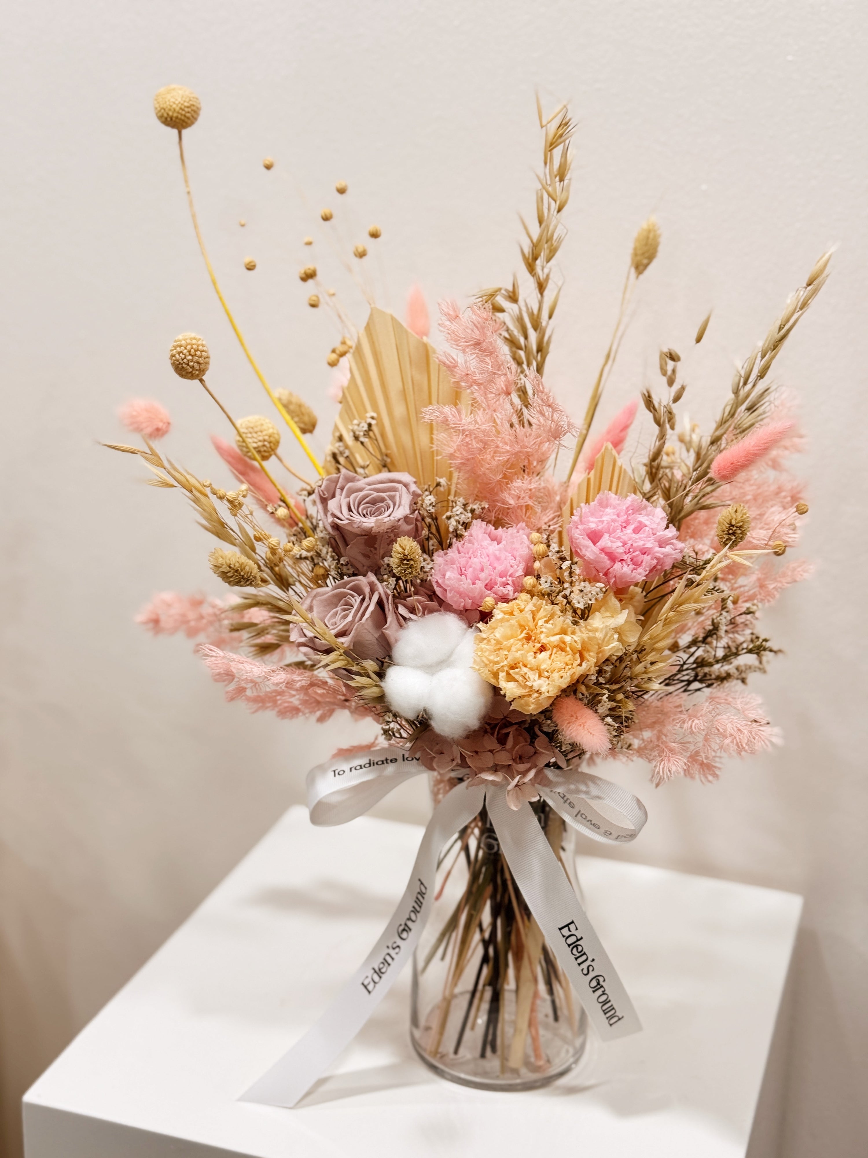 Soft Radiance, a preserved flower vase arrangement, featuring roses, carnations, fluffy cotton, and textural elements like bunny tails, grains, and palm spears, presented in a clear glass vase, crafted by Eden's Ground.