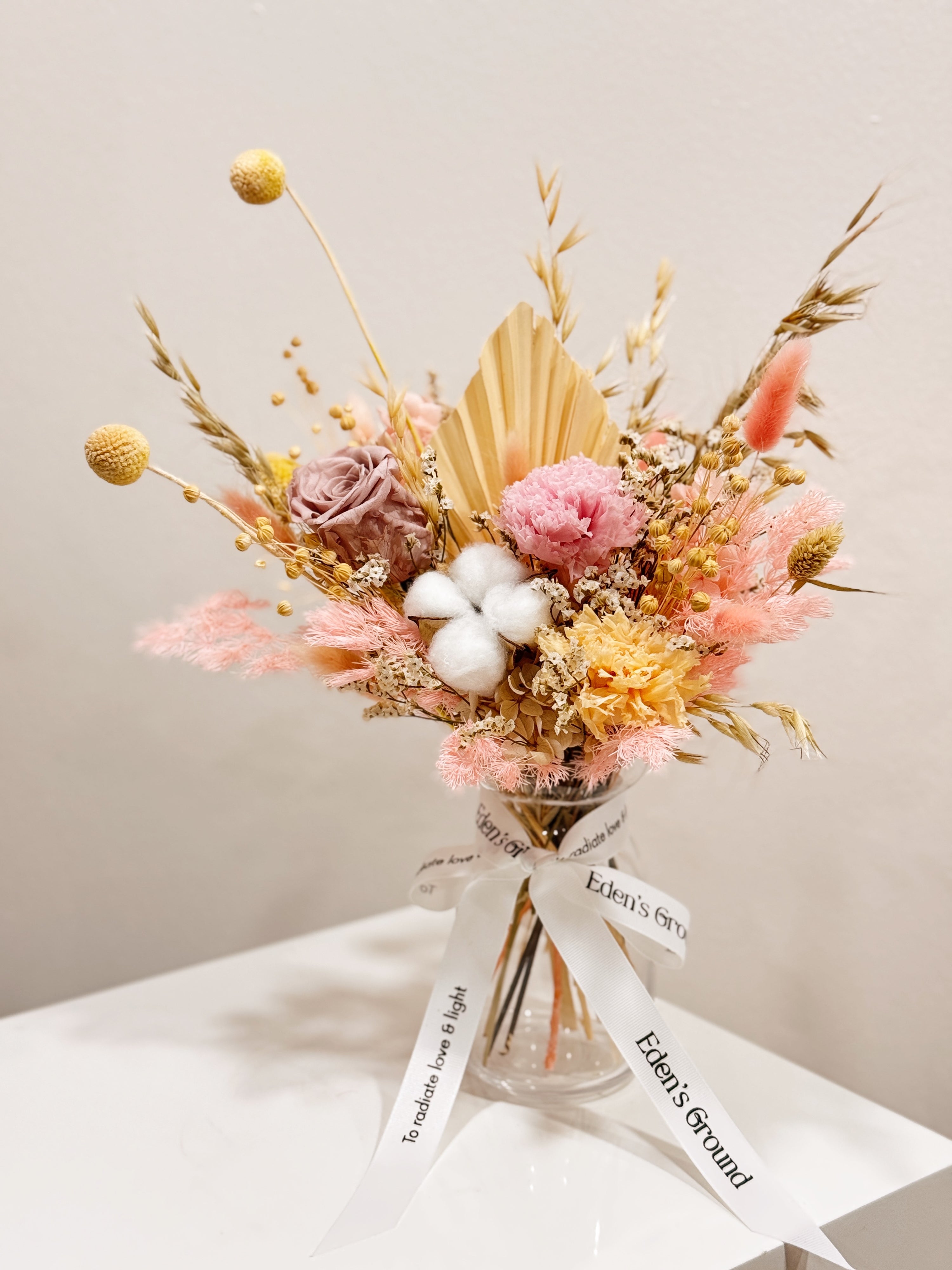 Soft Radiance, a preserved flower vase arrangement, featuring roses, carnations, fluffy cotton, and textural elements like bunny tails, grains, and palm spears, presented in a clear glass vase, crafted by Eden's Ground.