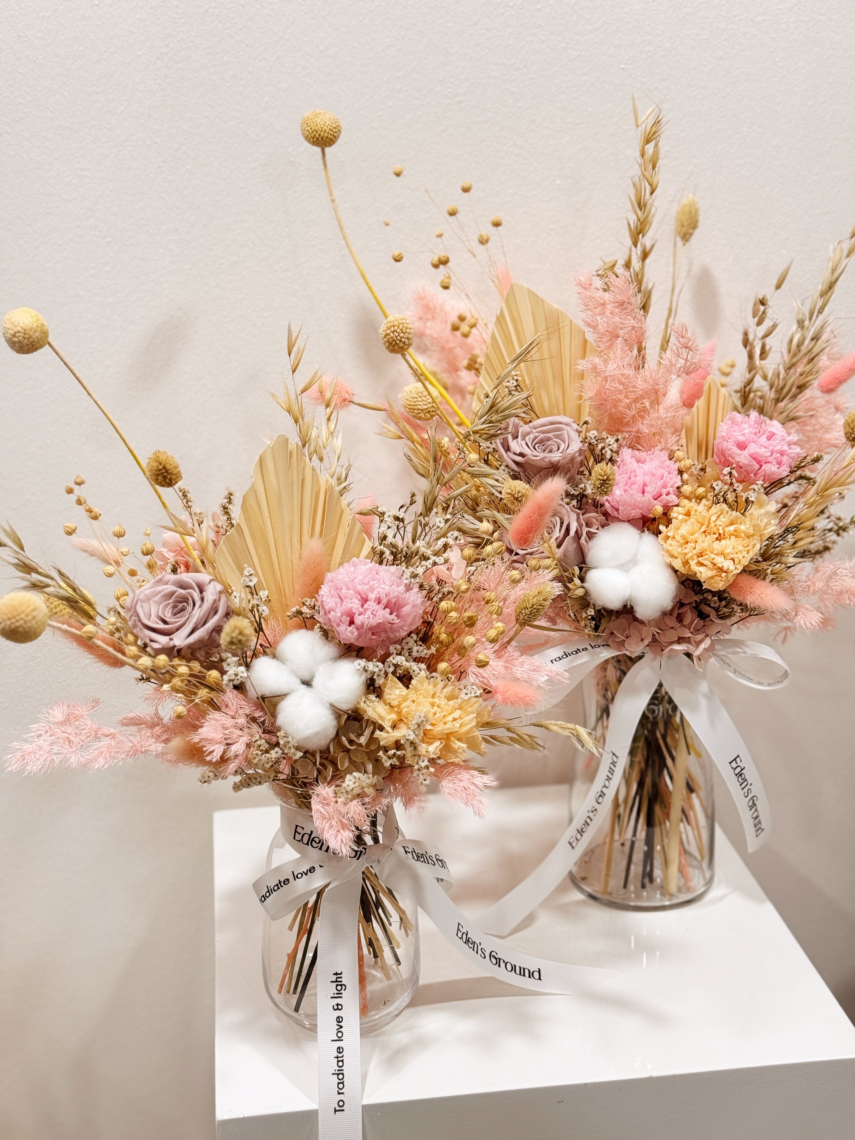 Soft Radiance, a preserved flower vase arrangement, featuring roses, carnations, fluffy cotton, and textural elements like bunny tails, grains, and palm spears, presented in a clear glass vase, crafted by Eden's Ground.