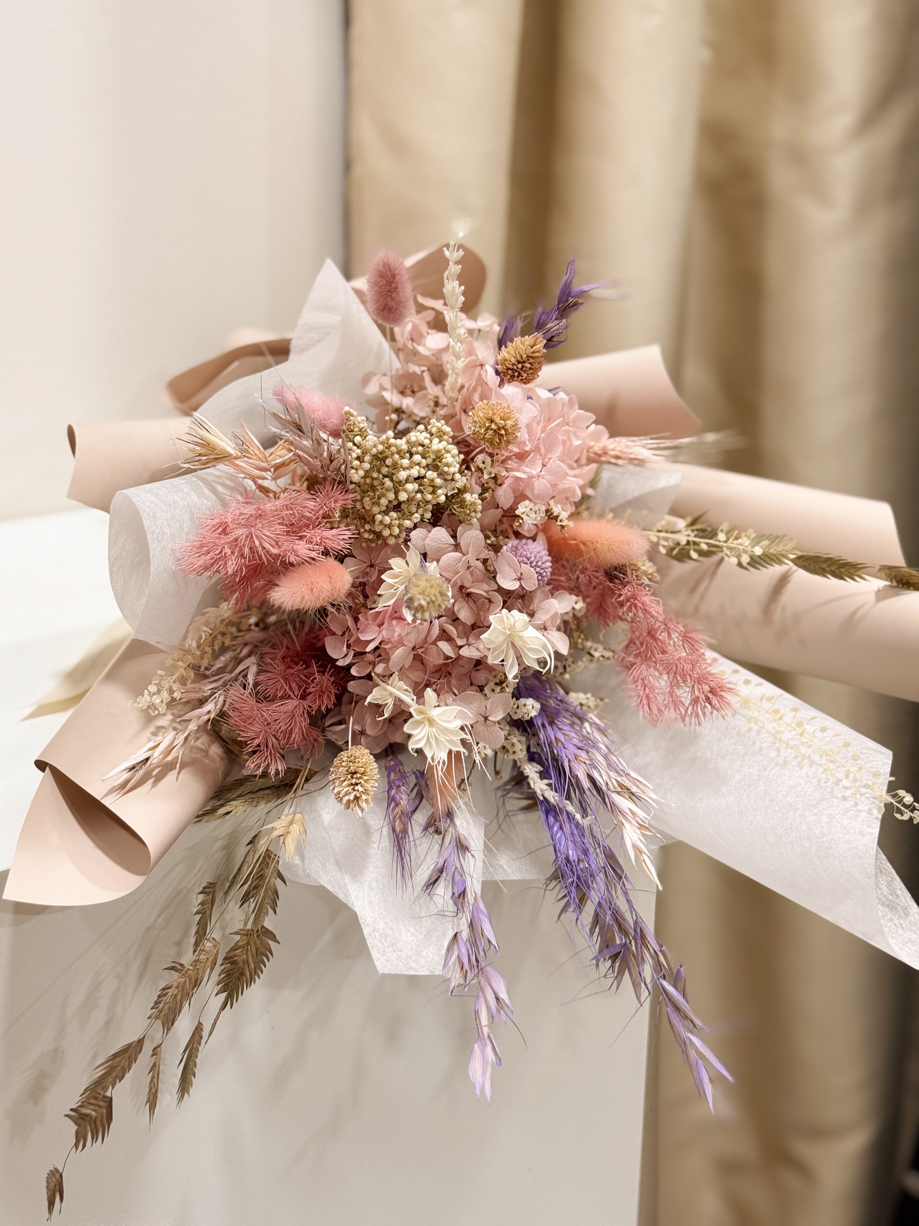 Still Blooming is a delicate preserved flower bouquet featuring soft pink and lavender blooms, thoughtfully gathered with textured foliage. Wrapped in white tissue and blush pink glassine, it is gently crafted by Eden’s Ground, a floral design boutique.